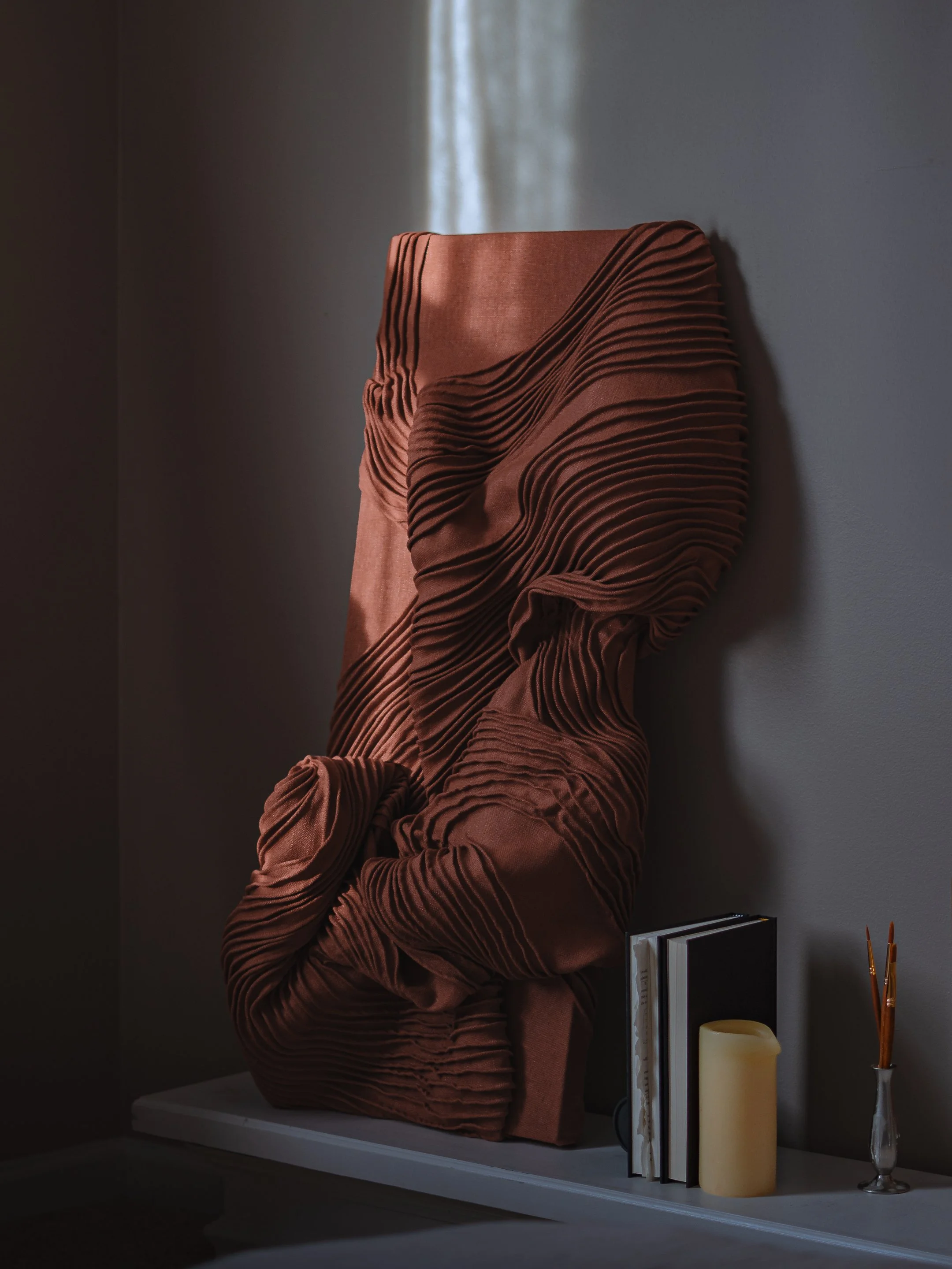 Decorative sculpture with layered, wavy, brown fabric-like design on a white surface, accompanied by books, a candle, and a small vase with paintbrushes.