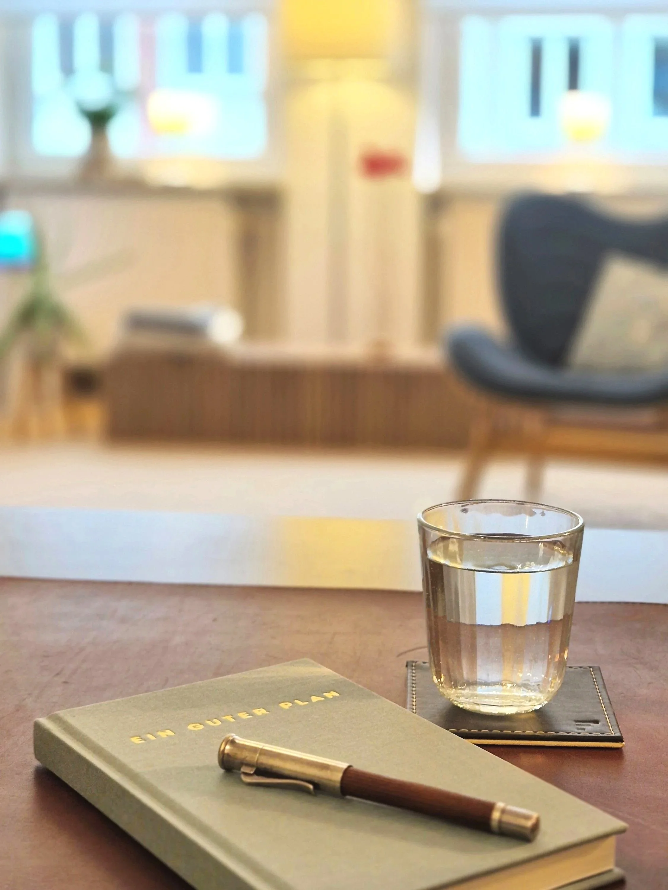 Im Vordergrund ein Buch mit der Aufschrift "ein guter Plan", ein Wasserglas, im Hintergrund ein freundlicher, heller Praxisraum