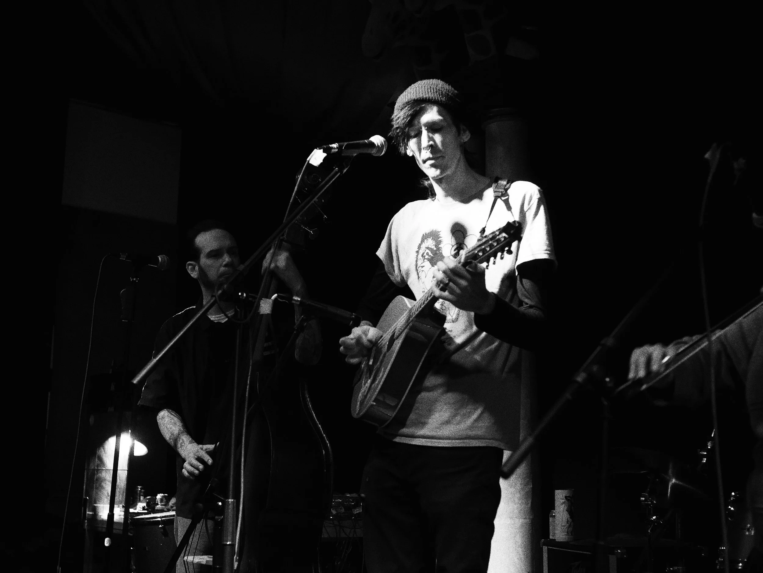 A young man with long hair, wearing a beanie and a T-shirt with a lion print, is playing an acoustic guitar and singing into a microphone on stage. Other band members and equipment are visible in the background.