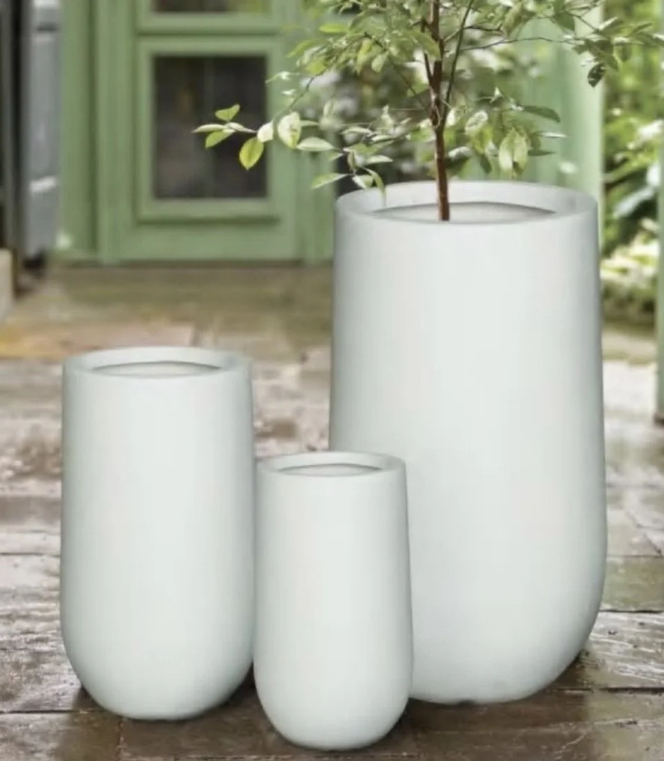 Three white modern vases of different heights on a wooden surface, with a potted plant in the tallest vase, in front of a green door.