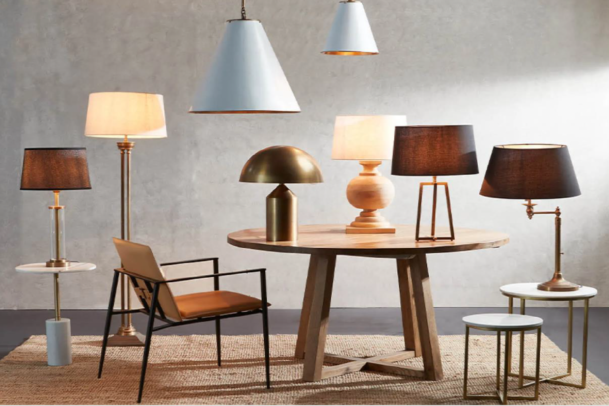 A modern living room setup featuring a round wooden table with two lamps, five standing lamps with different shades, a black chair with a tan cushion, and a beige rug.