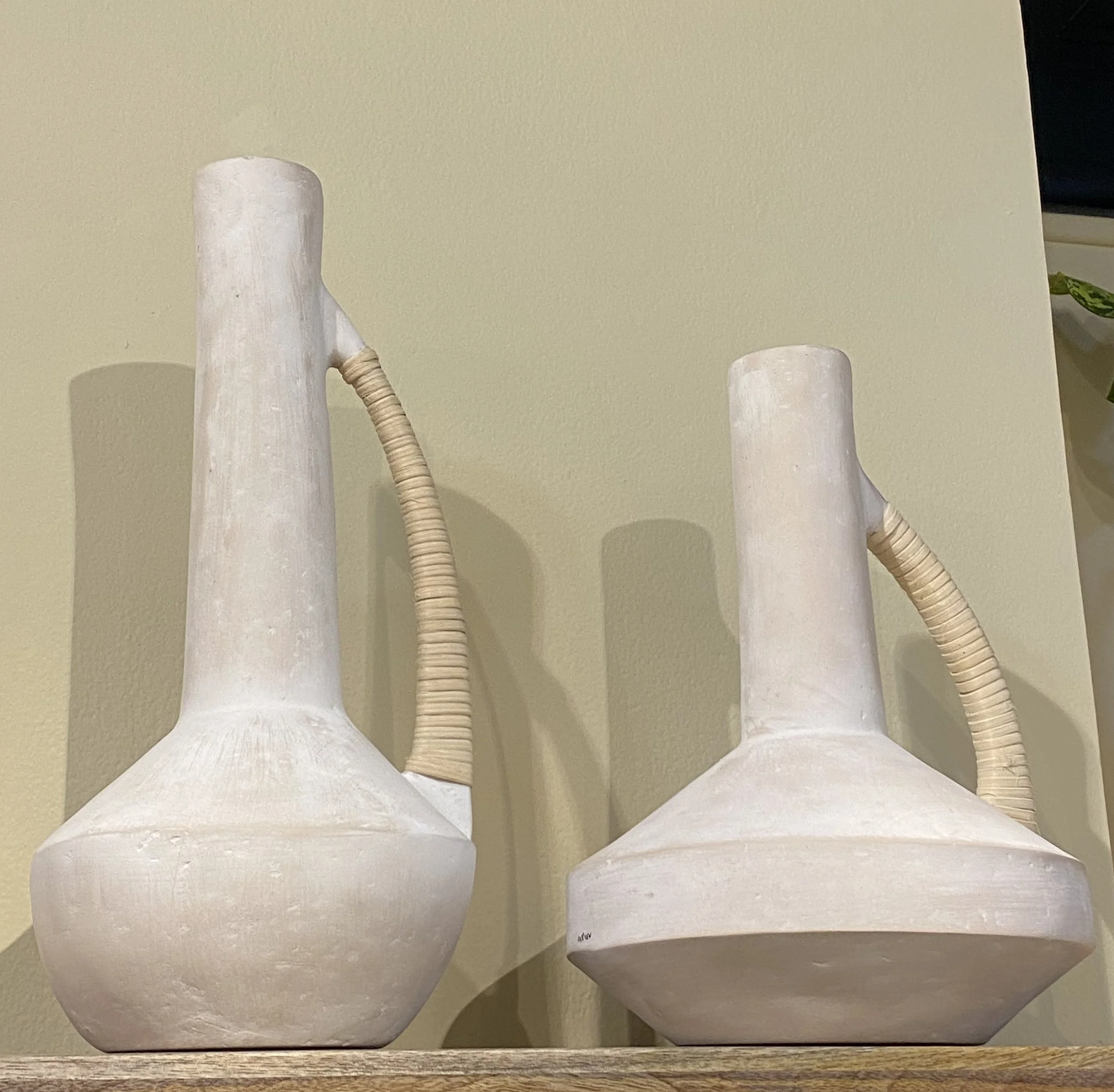 Two white ceramic vases with narrow necks and curved handles, placed on a wooden surface against a beige wall, casting shadows.