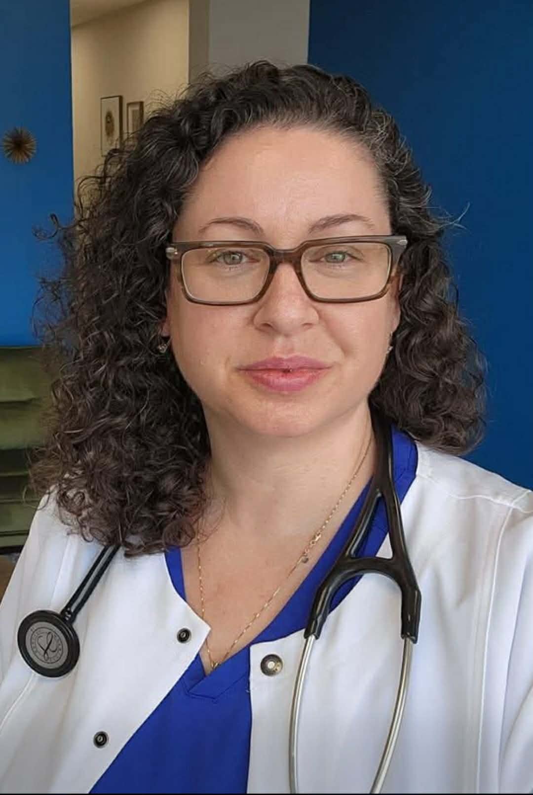 A woman with curly dark hair, wearing glasses, a white medical coat, and a blue top, smiling with a stethoscope around her neck.