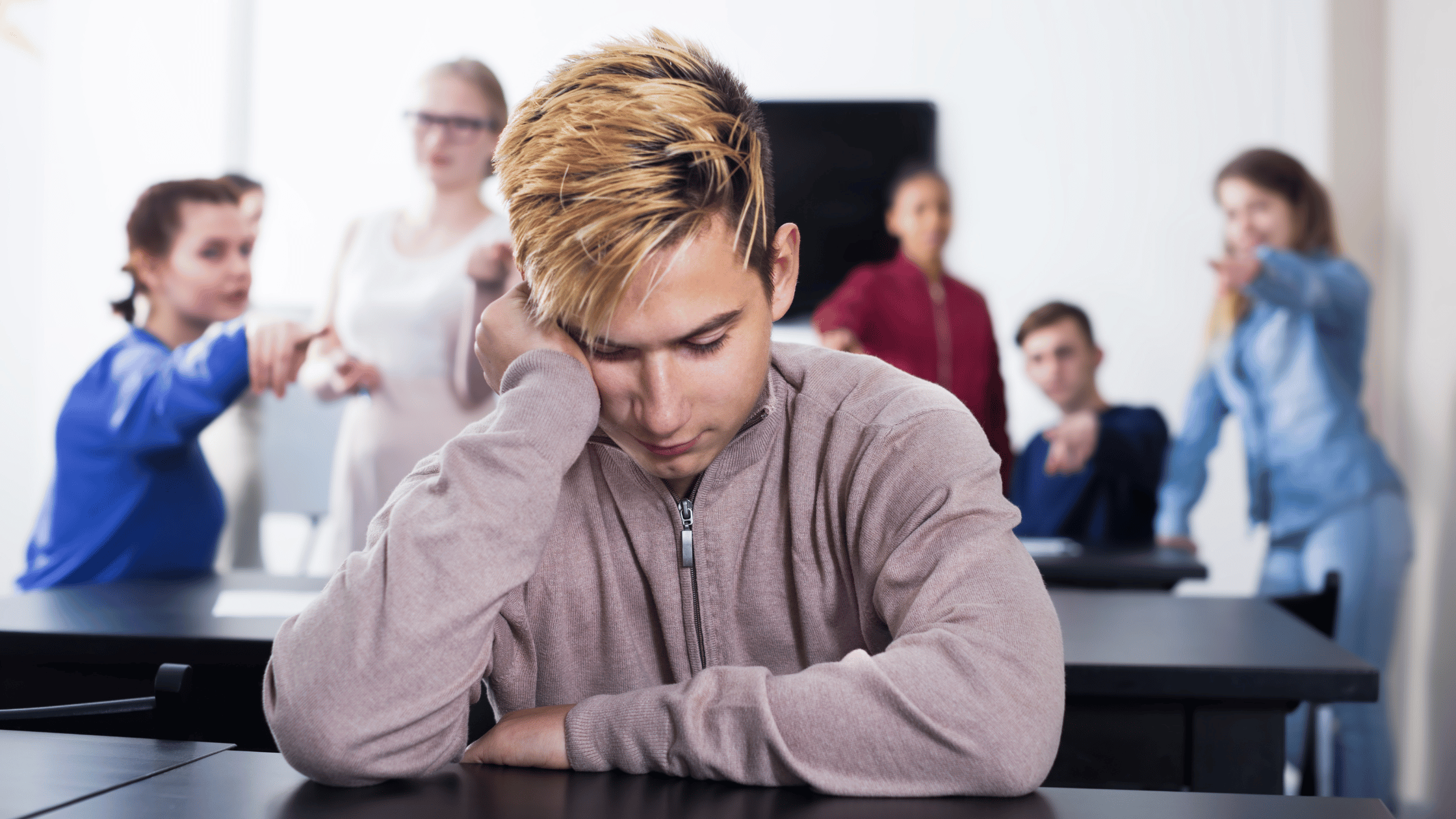 Male student with hand to cheek sitting at table alone with other students behind him points fingers at him.