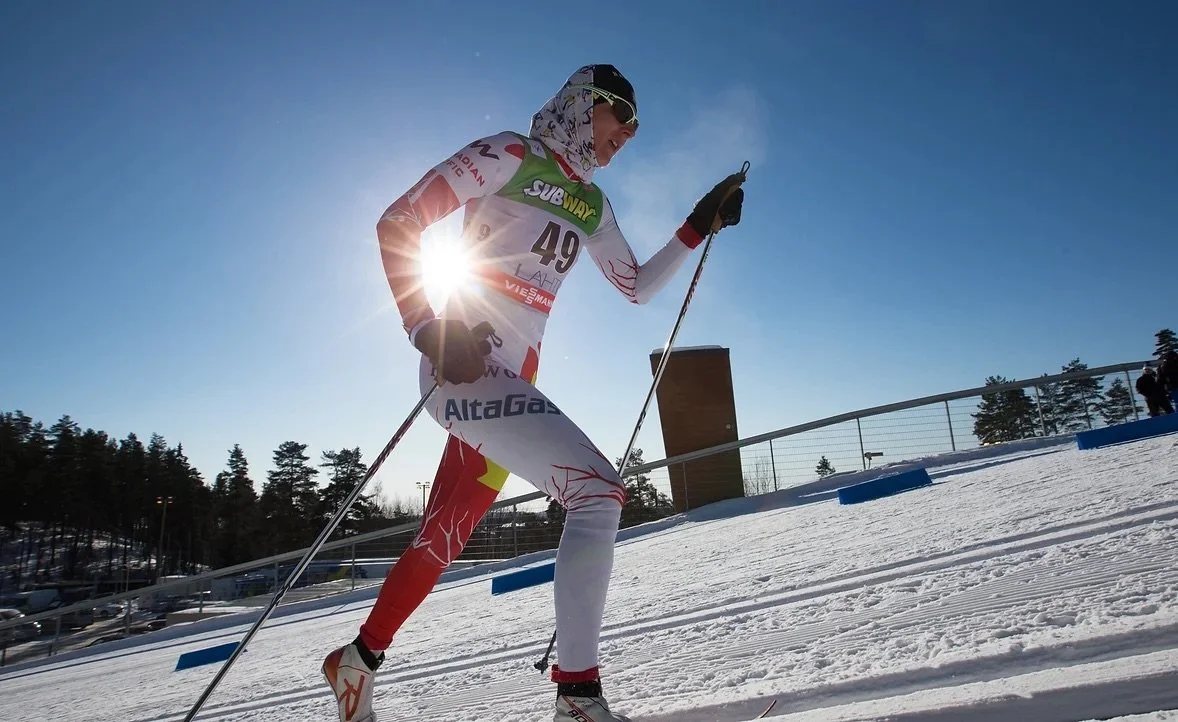 A cross-country skier in racing gear and sunglasses, marked with sponsor logos, skiing on snow with ski poles against a bright blue sky and sunlight