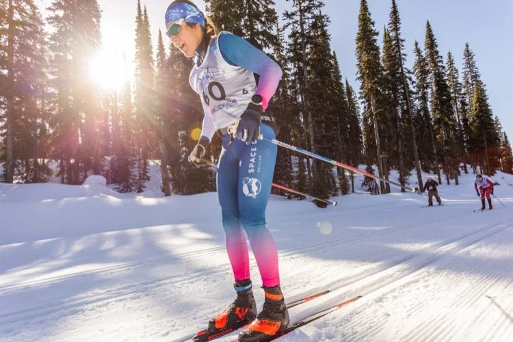 A woman in pink and red ski gear standing on snow with skis and poles, smiling with a mountain and blue sky in the background.