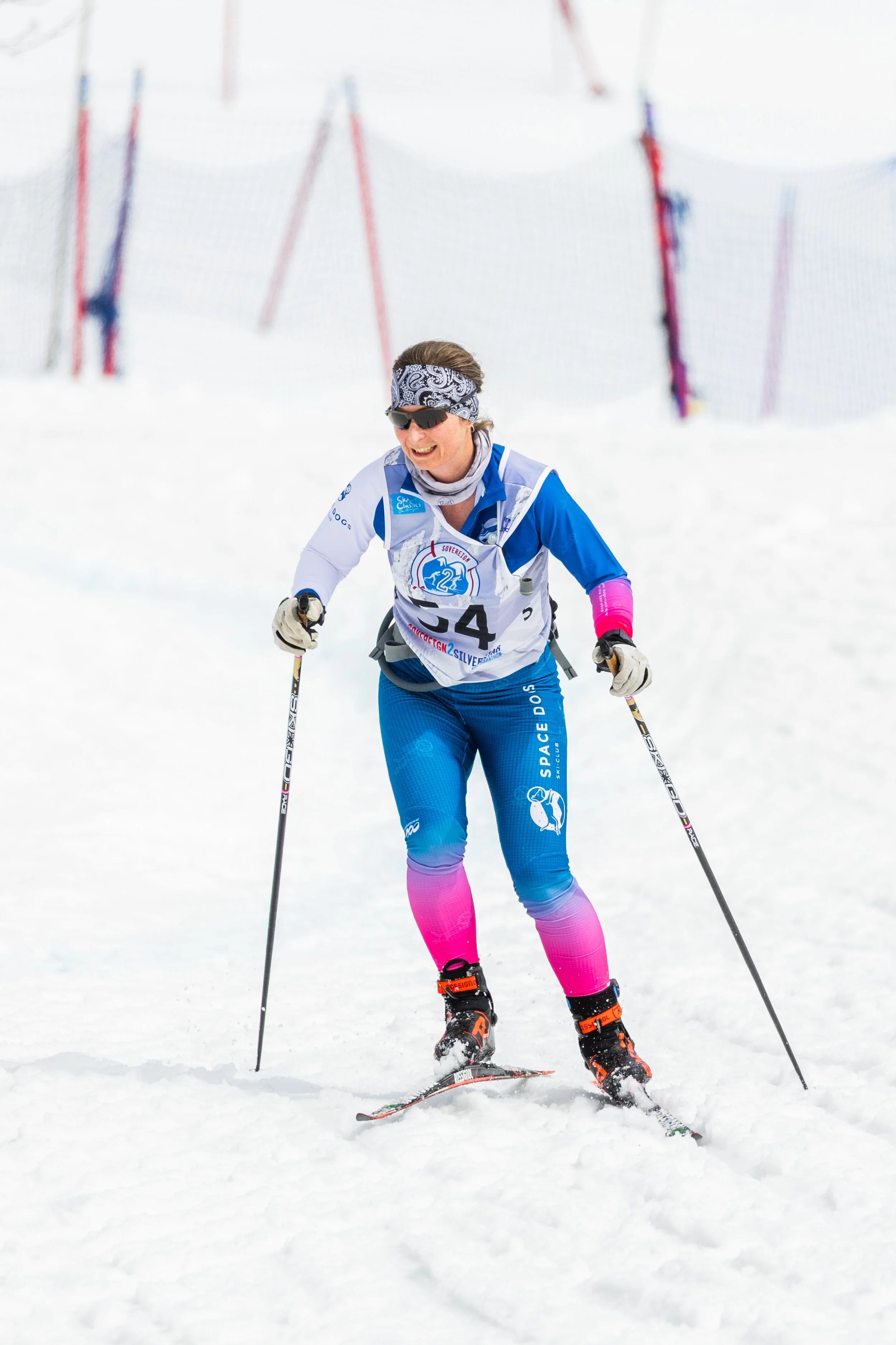 Woman wearing a blue jacket and a blue winter hat with goggles, smiling in a snowy forest landscape with snow-covered trees and overcast sky in the background.