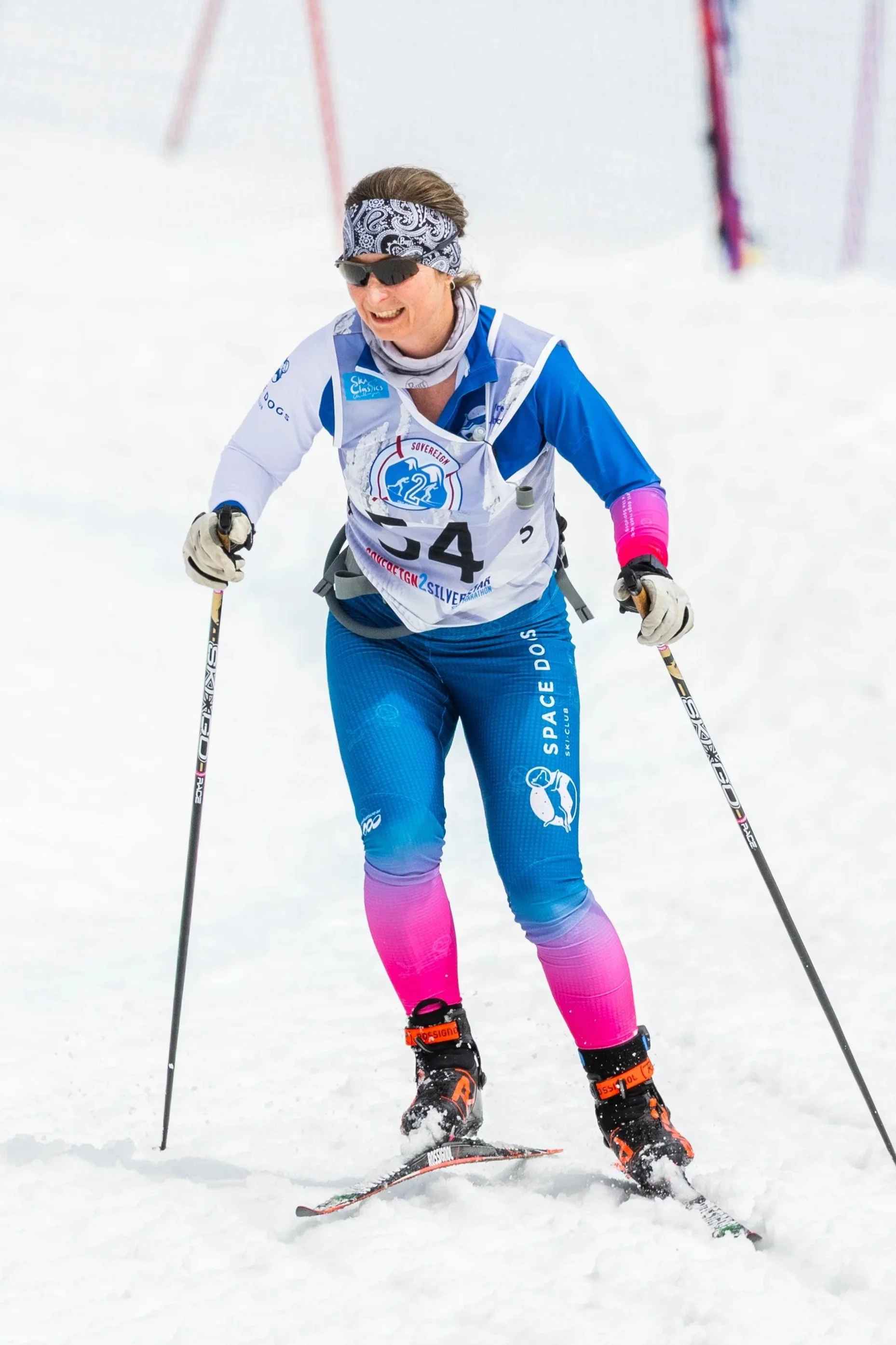 A woman dressed in athletic winter gear, holding a small container, standing on snow with skis, in a snowy mountainous forest.