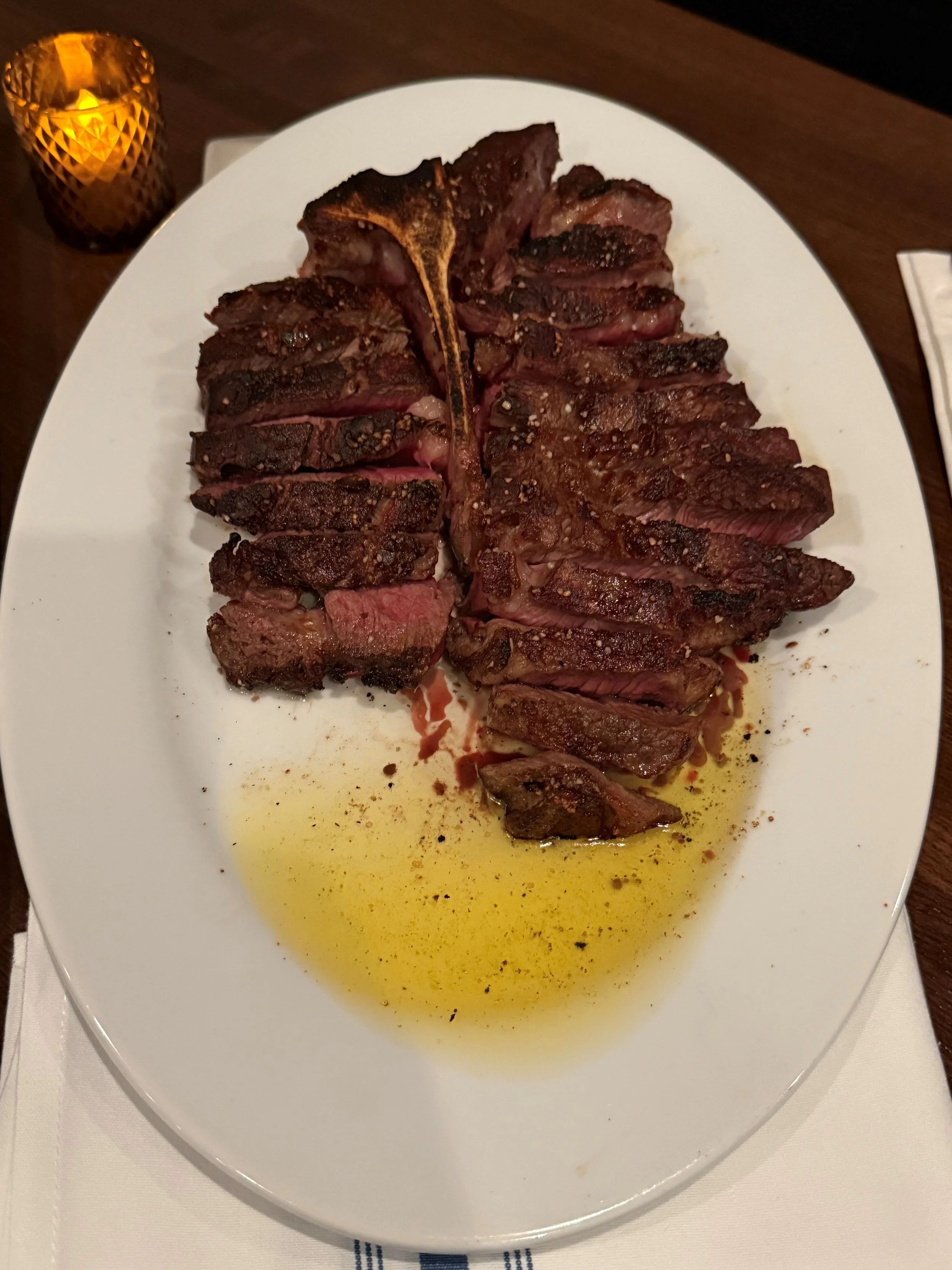 Porterhouse steak for two, served on a white oval plate with olive oil and black pepper, on a dark wooden table.