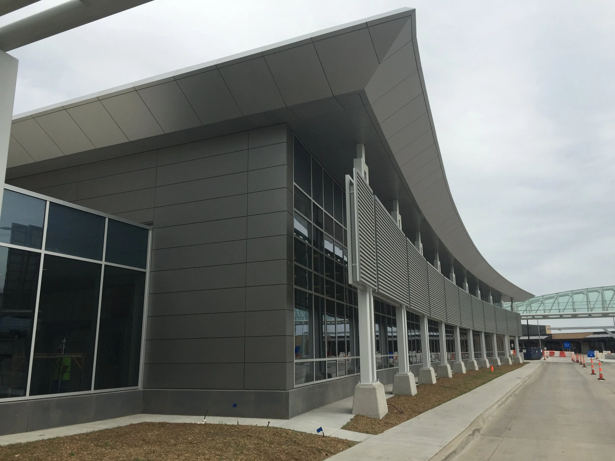 Modern airport terminal building with curved roof, large glass windows, and an exterior walkway with orange traffic cones.