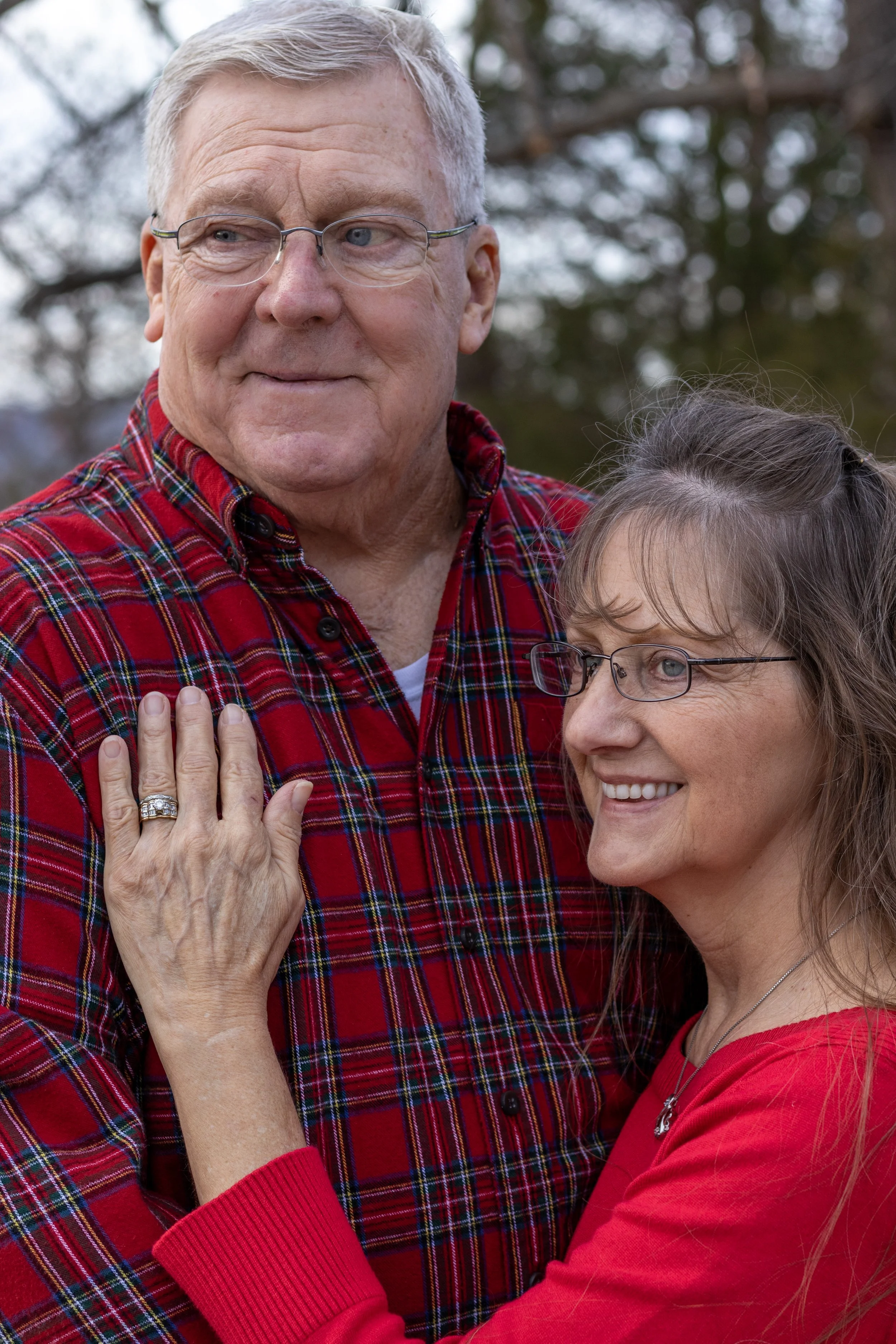 An elderly man and woman wearing glasses and red clothing, standing outdoors, smiling and embracing each other.