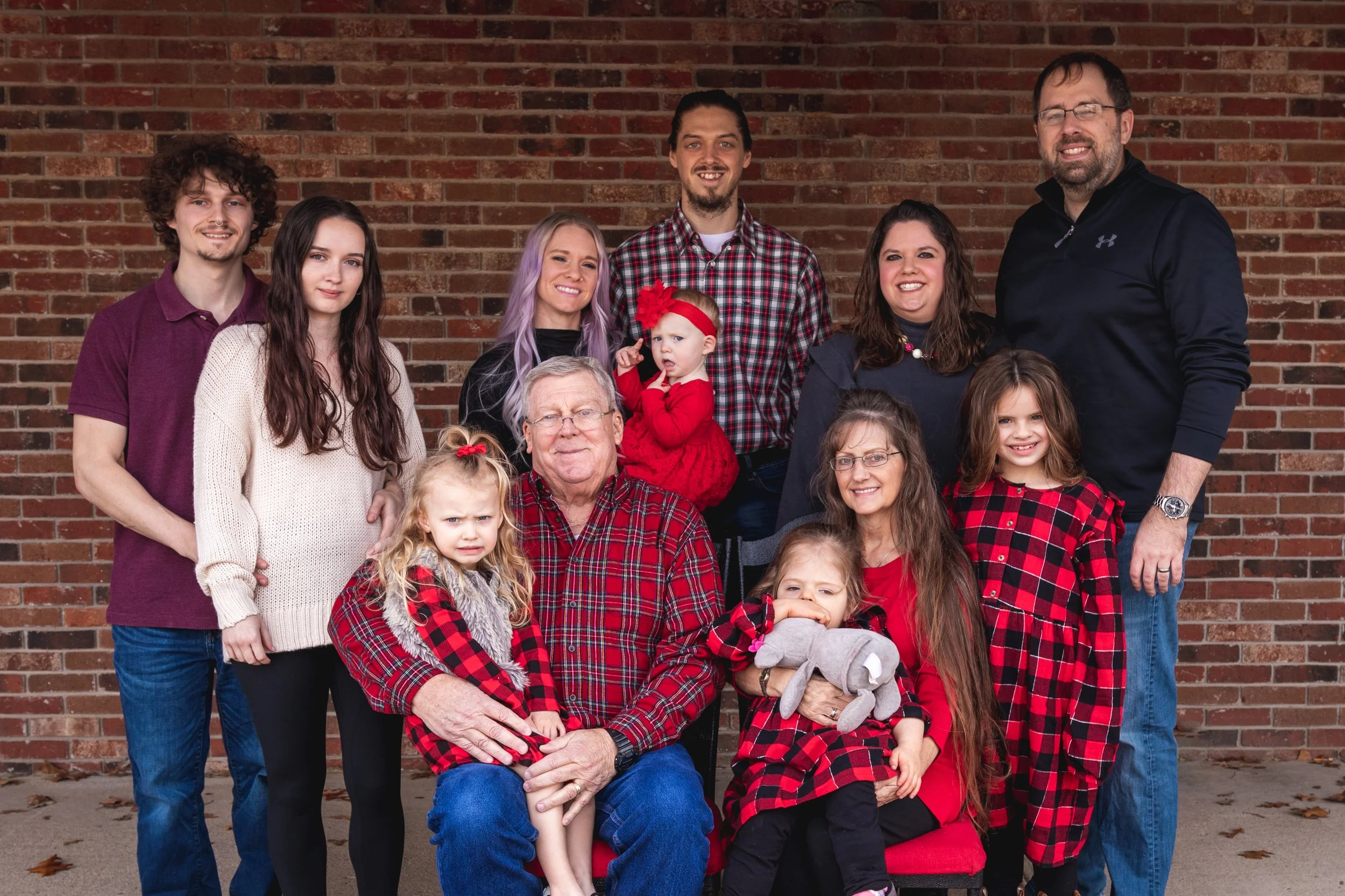 Family photo of multiple generations in front of a brick wall, wearing red, black, purple, and cream clothing, with some children holding stuffed animals.