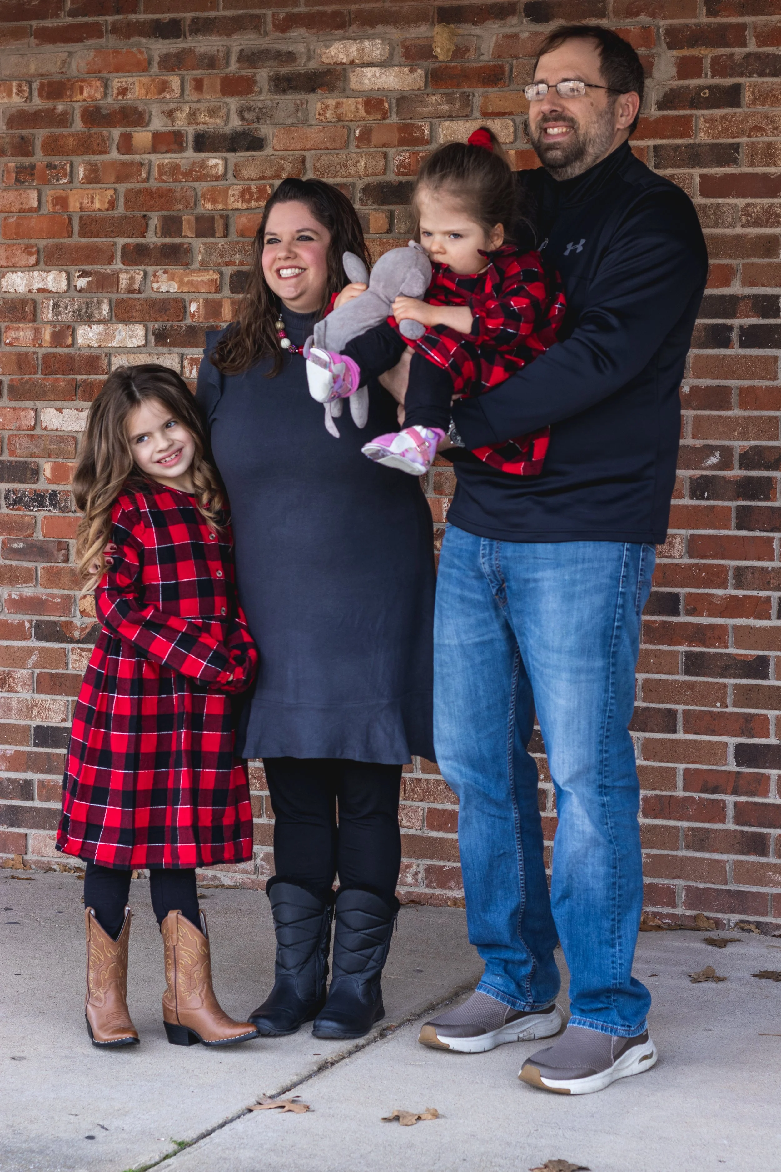 Family of four standing outside against a brick wall, with two young girls in red and black plaid dresses, a woman in a dark dress, and a man holding a toddler girl in matching dress and taking a playful photo.