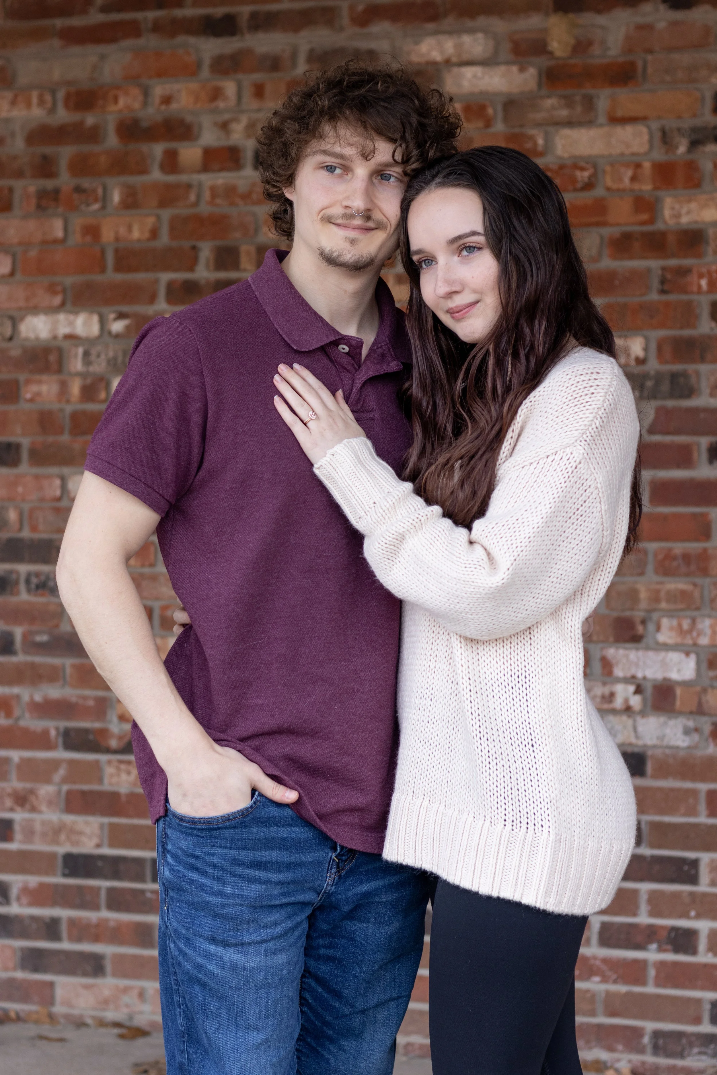 A young couple standing close together in front of a brick wall; the woman is resting her hand on the man's chest, showing her engagement ring.