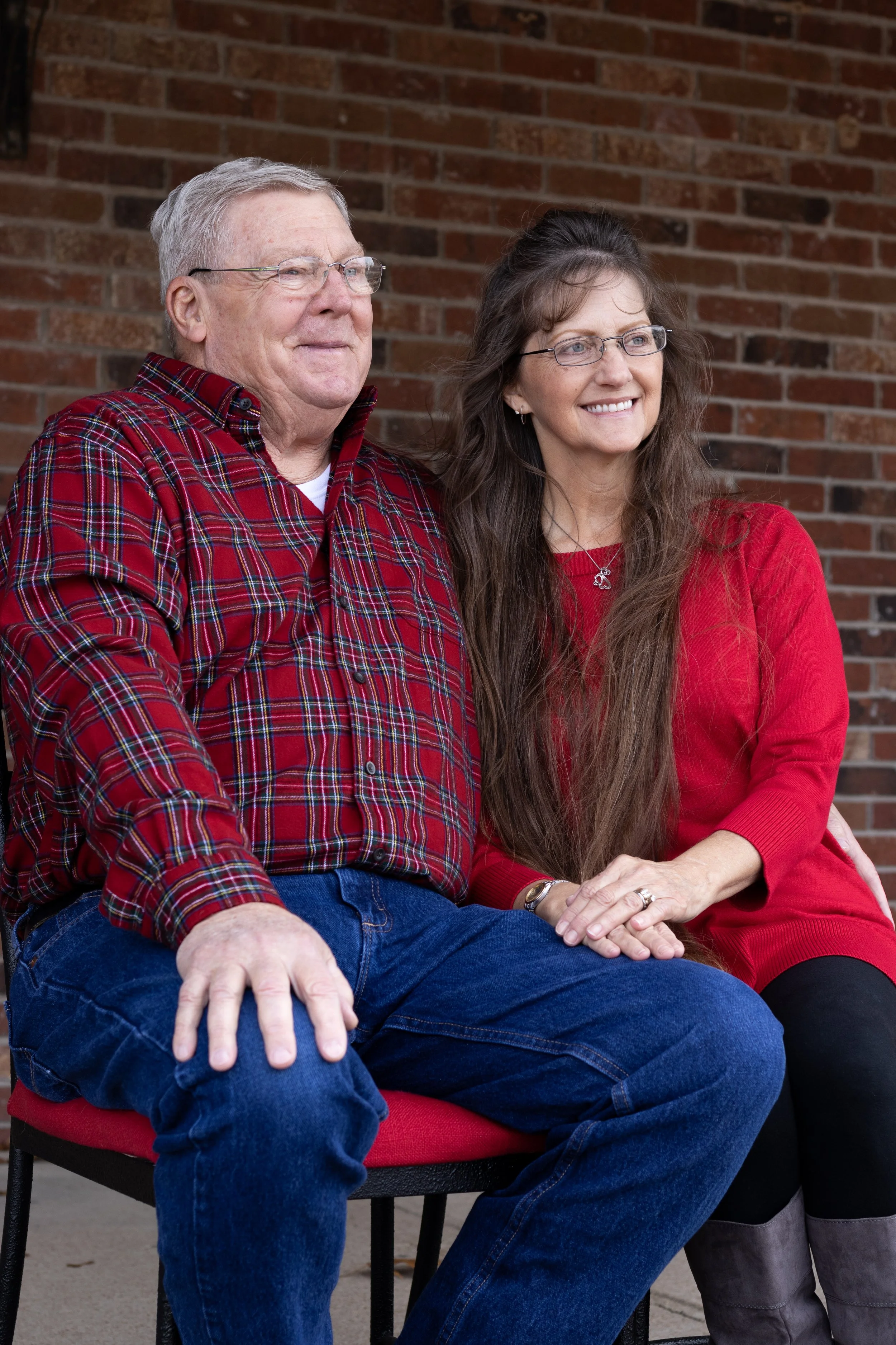 A happy elderly man and a middle-aged woman sitting close together in front of a brick wall, smiling.