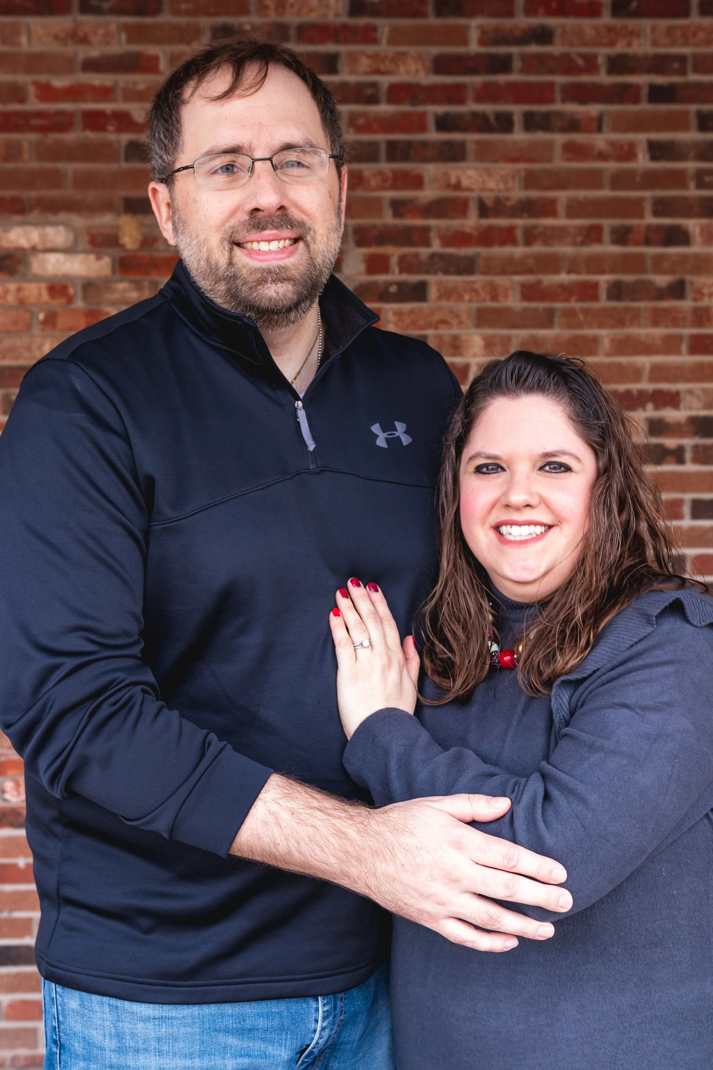 A smiling couple stands close together in front of a brick wall. The man is wearing glasses, a black Under Armour shirt, and has facial hair. The woman has long brown hair, red nail polish, and is wearing a dark gray jacket and a red necklace, with her hand on the man's chest showing a wedding ring.