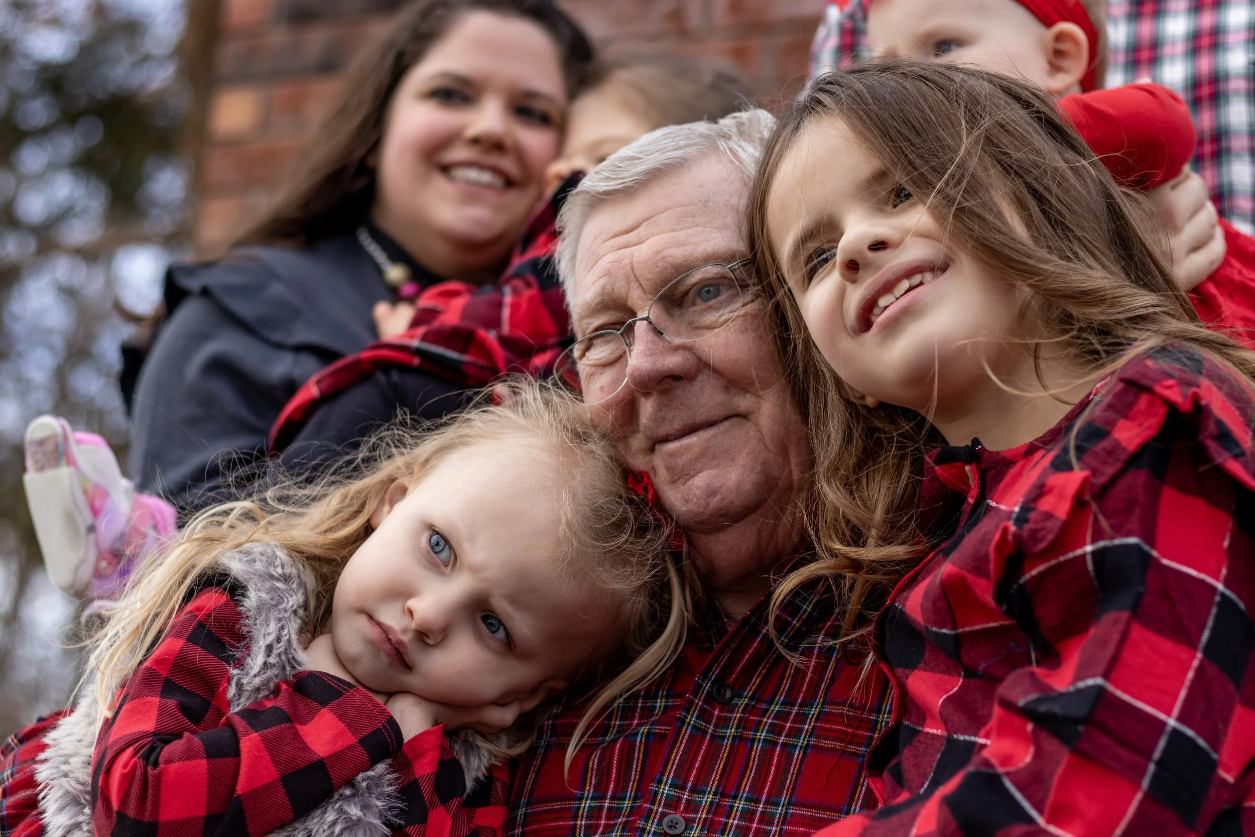 A family of six, including children and an elderly man, dressed in red and black plaid shirts, taking a close-up group selfie outdoors. They are smiling and embracing each other.