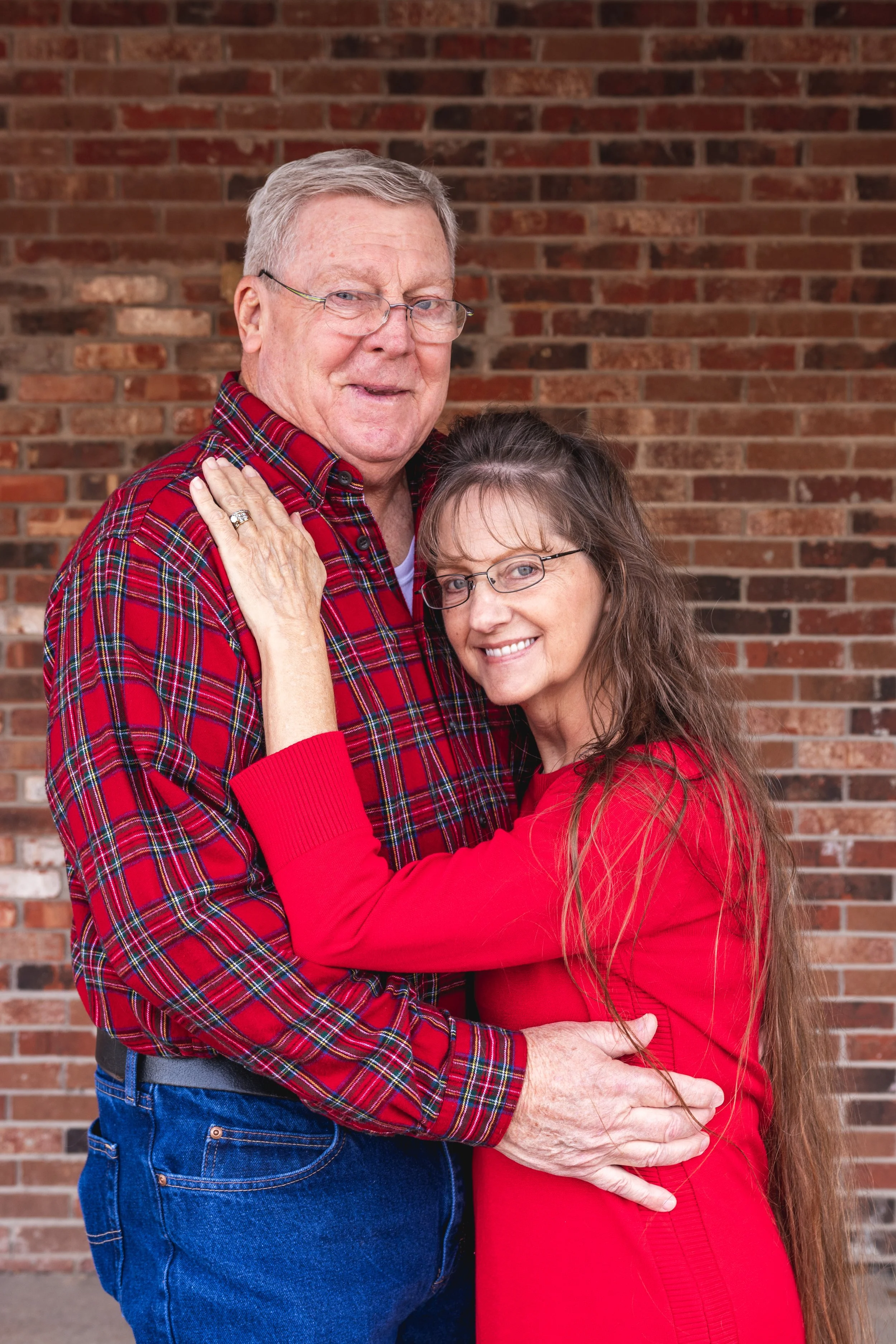 An elderly couple wearing red clothing, standing close together in front of a brick wall, smiling and embracing.