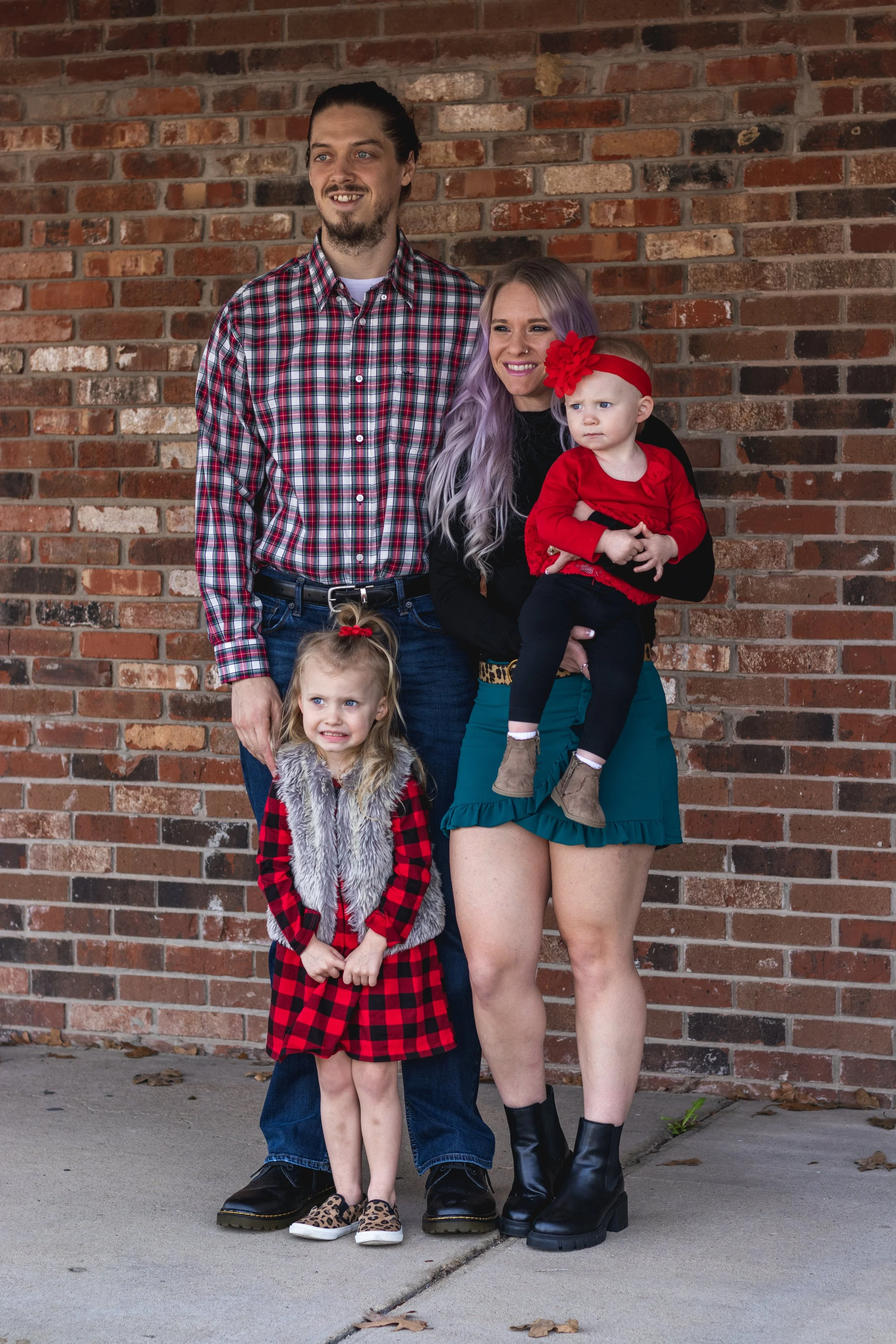 Family press photo against a brick wall, featuring a man, woman, and two young girls, all dressed in red and black with festive accessories.