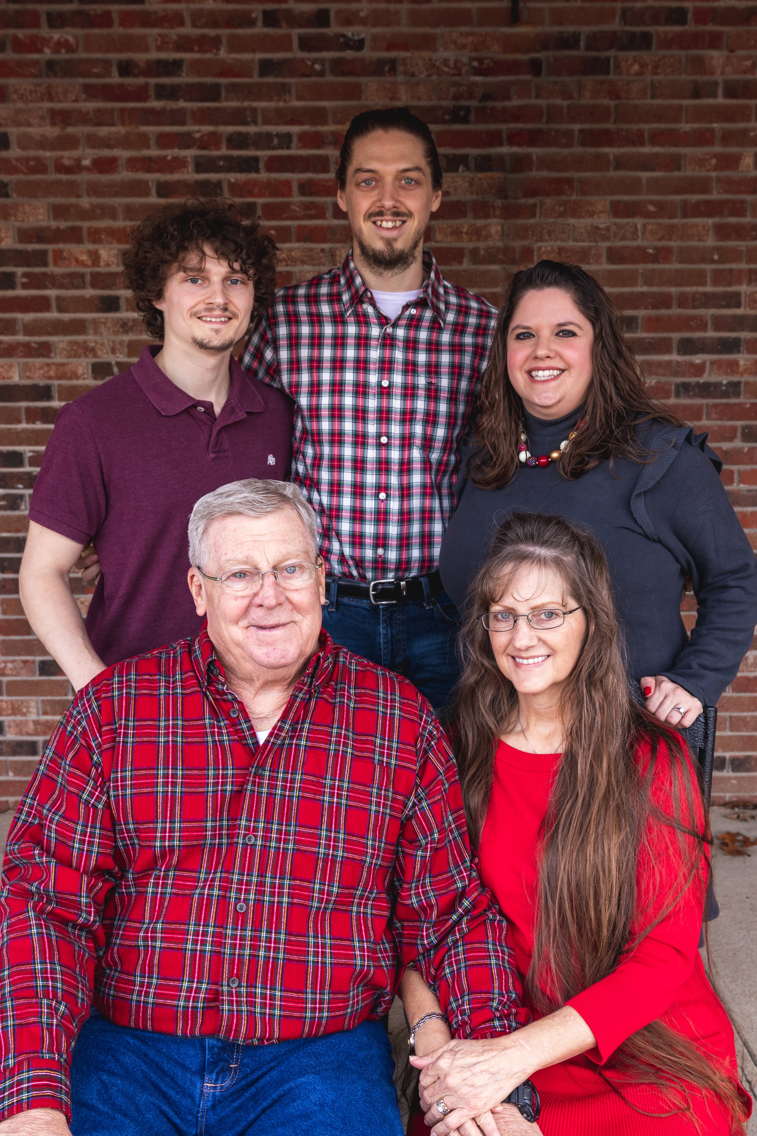 A multi-generational family photo of six people standing and sitting in front of a red brick wall, dressed in casual attire with some wearing plaid shirts, smiling at the camera.