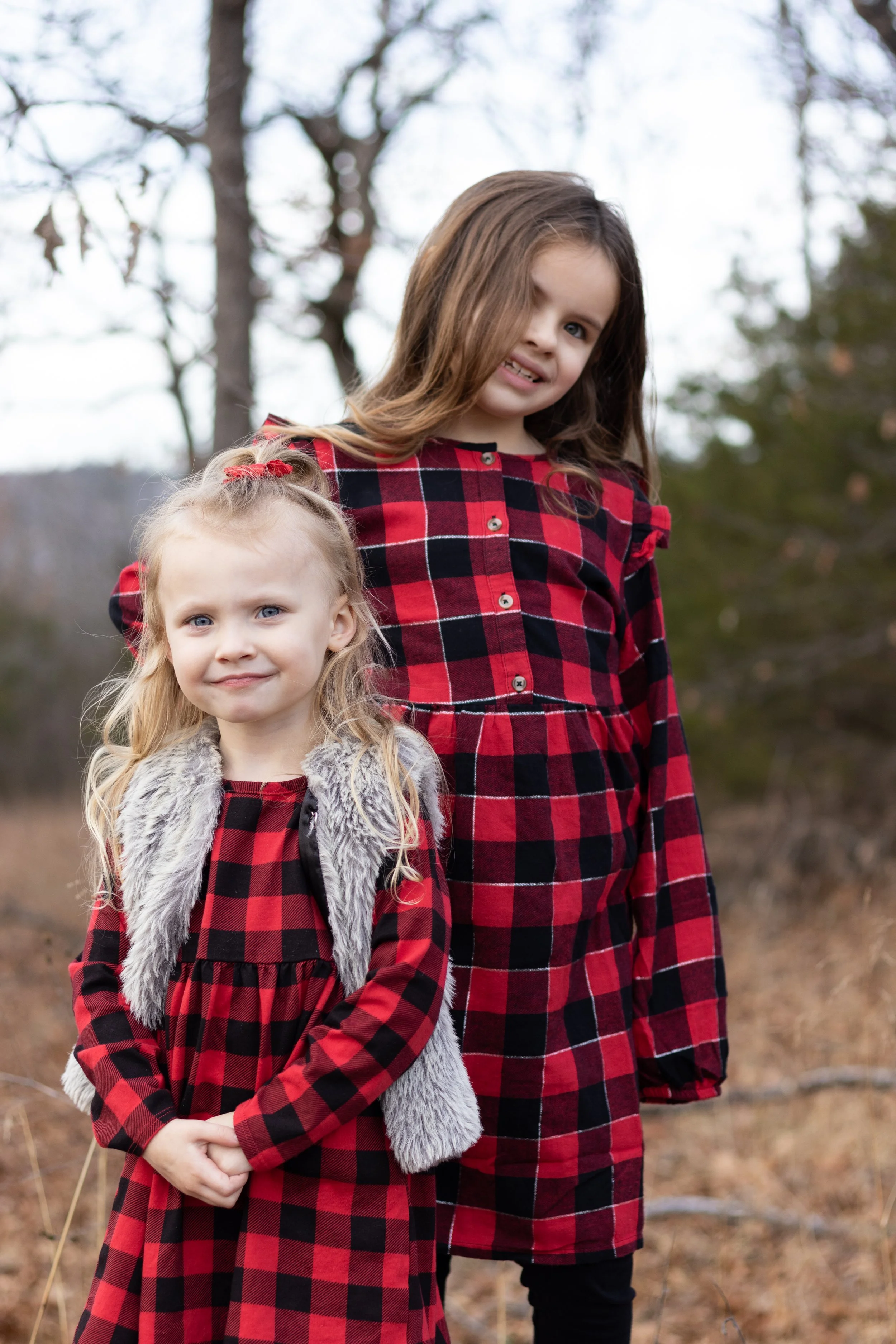 Two young girls outdoors in fall clothing, standing in a field with trees in the background, both wearing red and black plaid dresses.