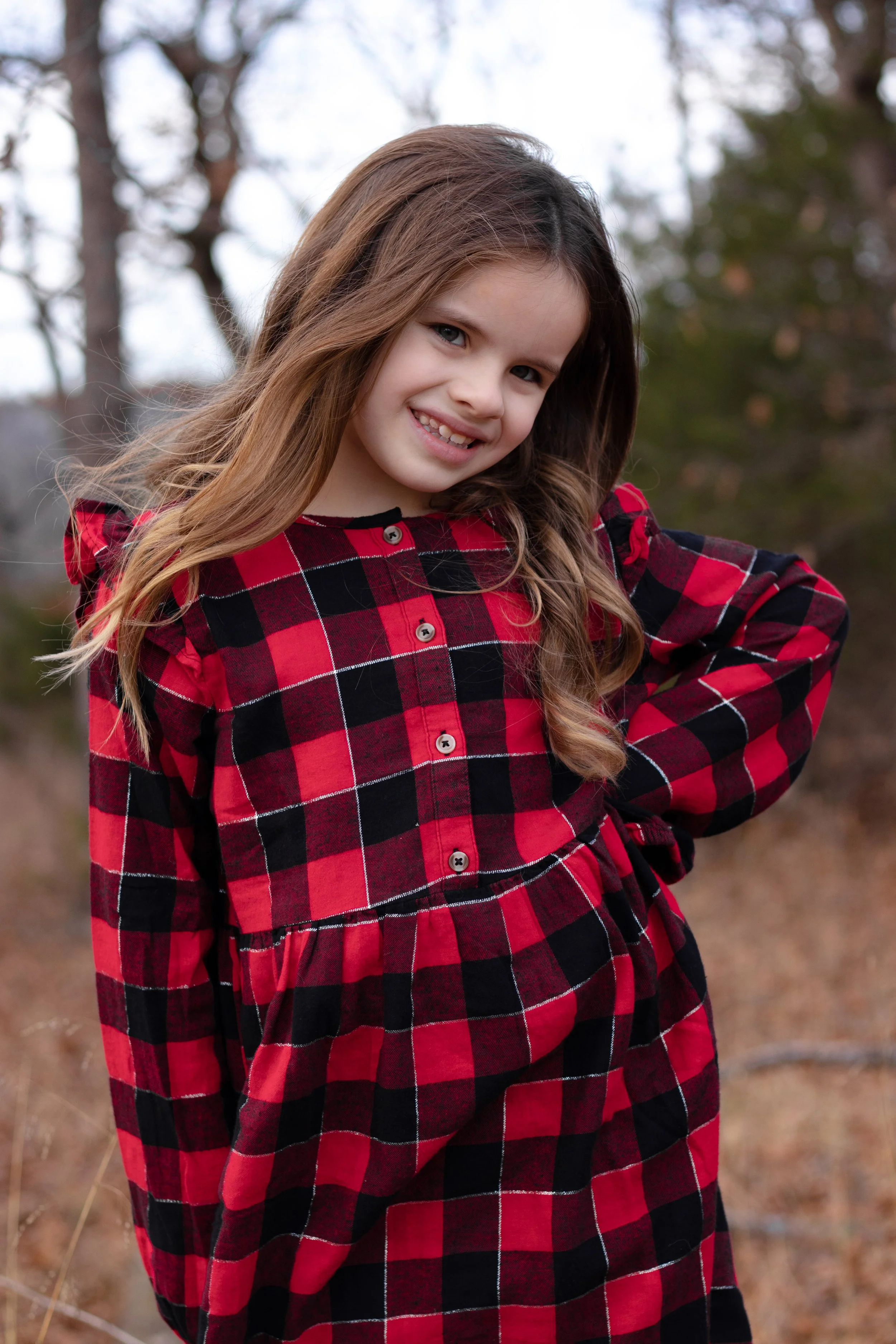 A young girl with long brown hair wearing a red and black plaid dress, standing outdoors with trees in the background, smiling at the camera.
