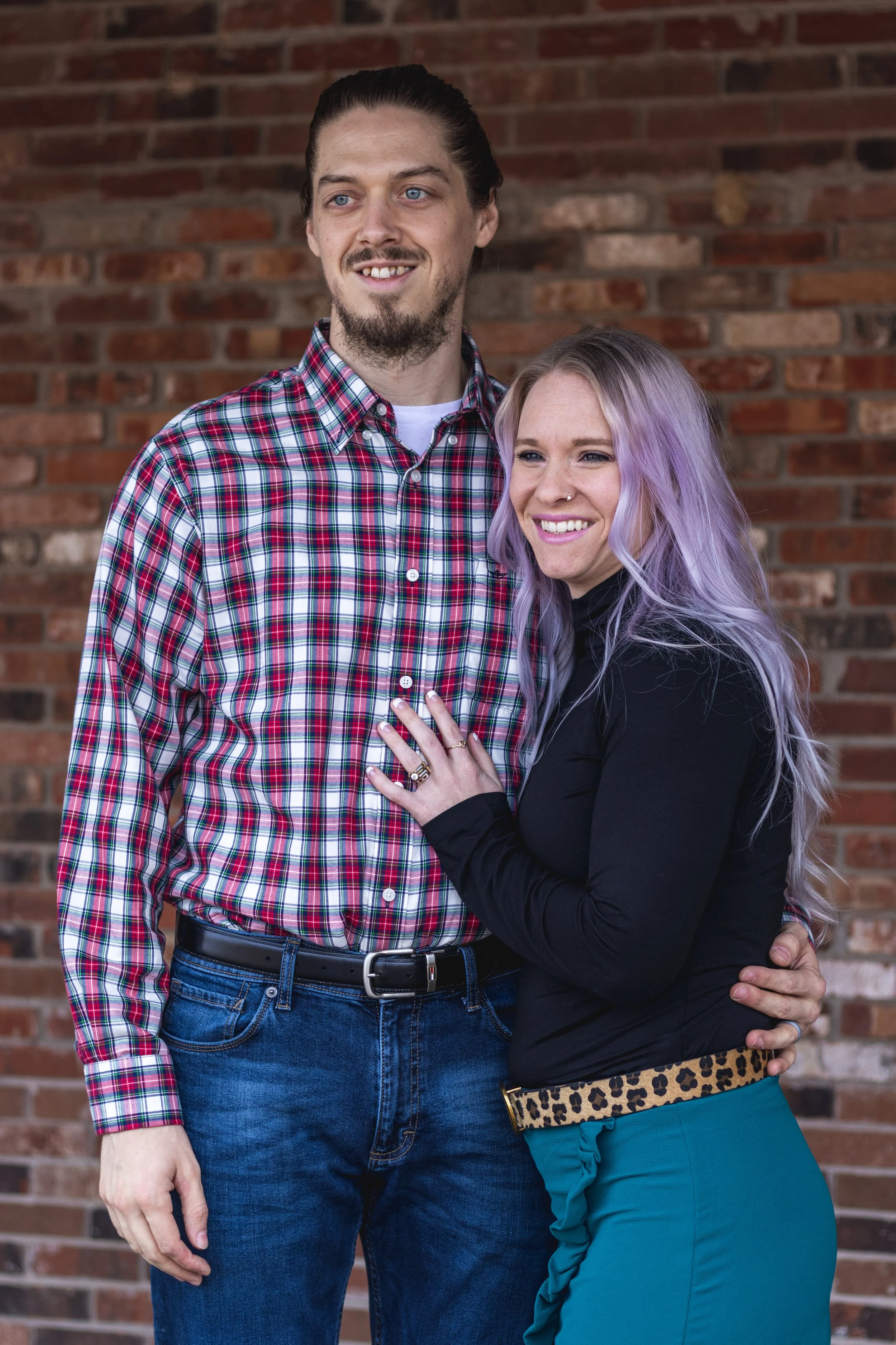 A man with a beard and mustache wearing a red plaid shirt and jeans, standing next to a woman with long, wavy, lavender hair, wearing a black top and teal pants, against a brick wall.