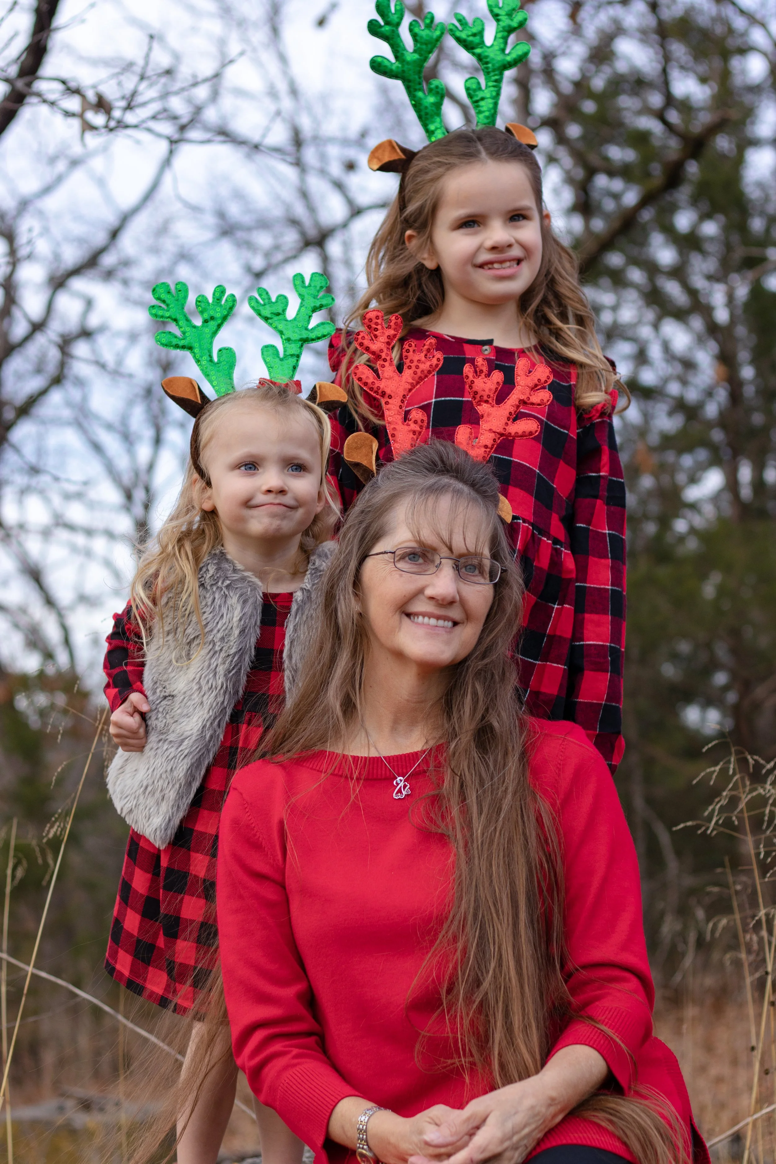 A woman and two girls outdoors, wearing festive reindeer antlers headbands, in a natural setting with trees, dressed in red and black plaid and red clothing, celebrating Christmas.