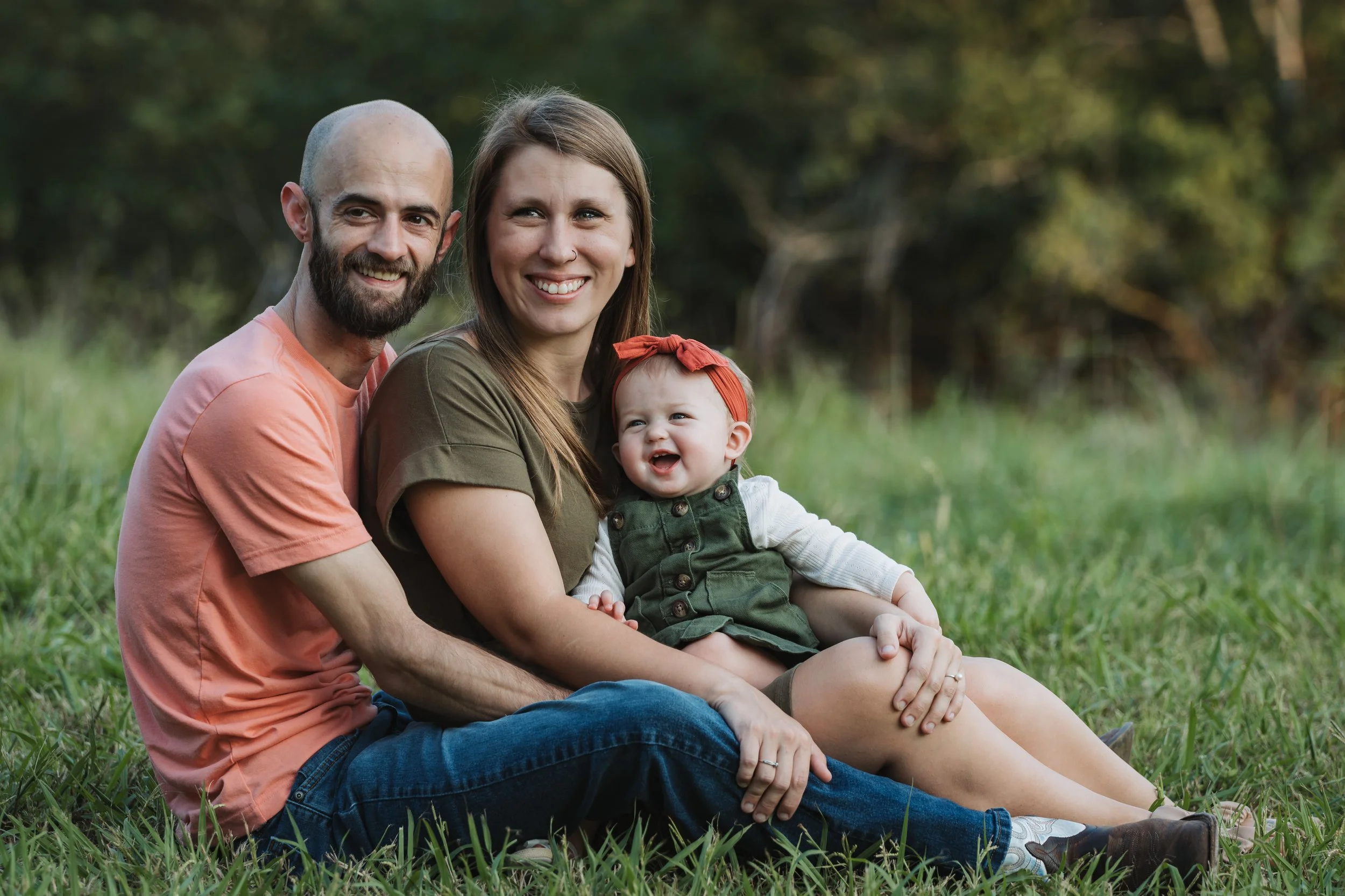 Family sitting on grass in a park, smiling, with trees in the background.