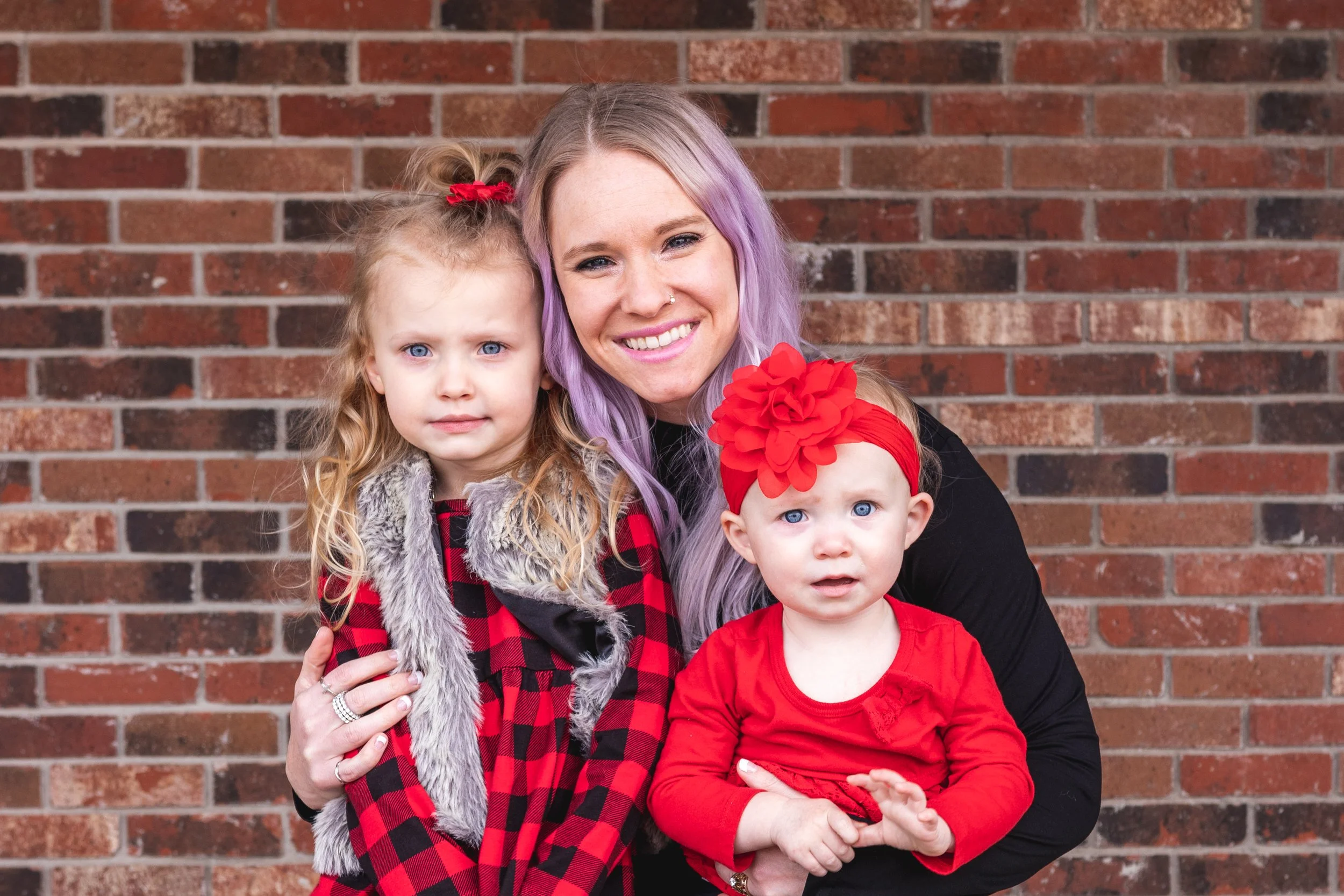 A woman with purple hair and two young girls, one with blonde hair and a red headband, and another with blonde hair and a red plaid dress, all standing in front of a brick wall.