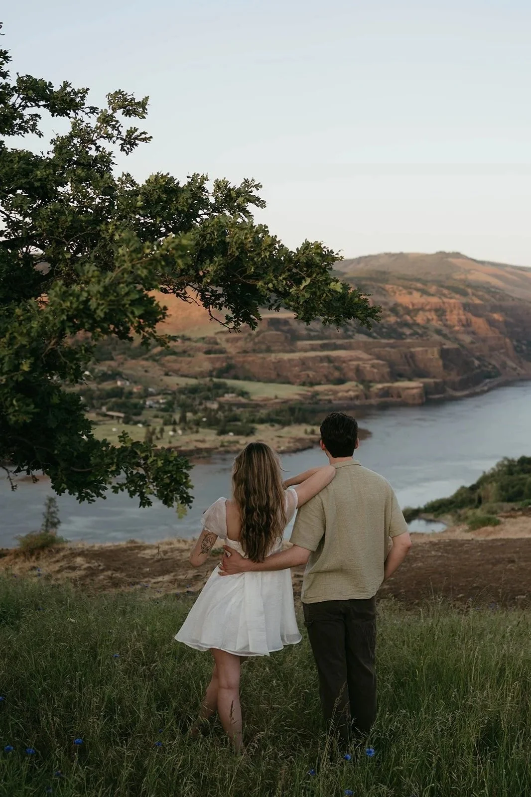 A couple stands on a grassy hill overlooking a river and rolling hills at sunset, with one person's arm around the other's shoulder.