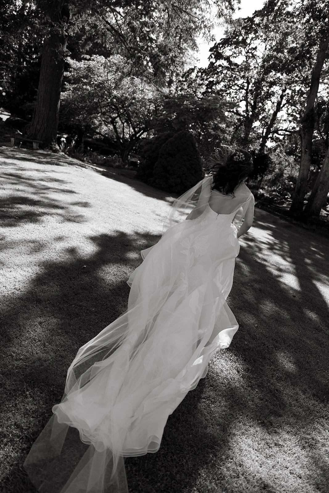 A woman in a wedding dress walking through a park with trees and bushes, sunlight casting shadows on the ground.