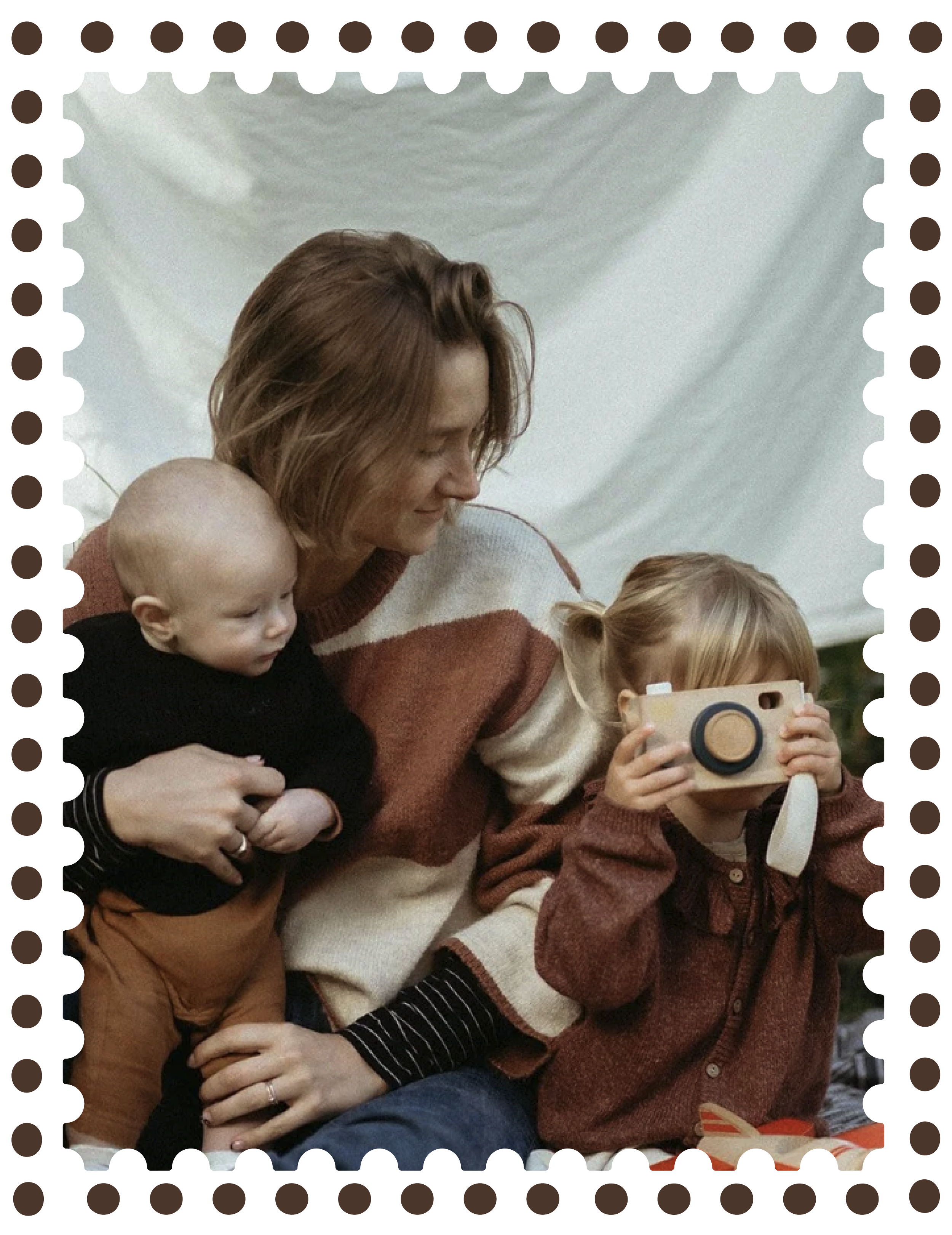 A woman with shoulder-length brown hair, wearing a striped black and white shirt, is holding a young child with blonde hair and a black shirt. Another young girl with blonde hair, wearing a reddish-brown sweater, is sitting in front of them, holding a camera, with a white scarf around her neck. They are sitting in front of a white backdrop or tent, engaging with a camera or device.