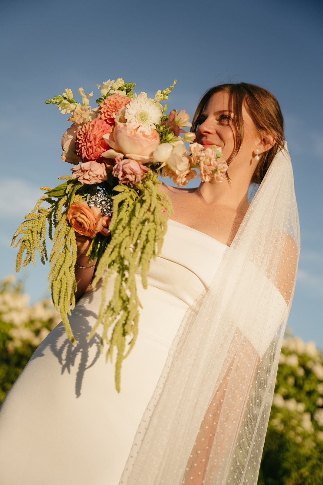 Brunette bride holding a large bouquet of pink and white flowers outside against a blue sky, wearing a white wedding dress with a sheer veil.
