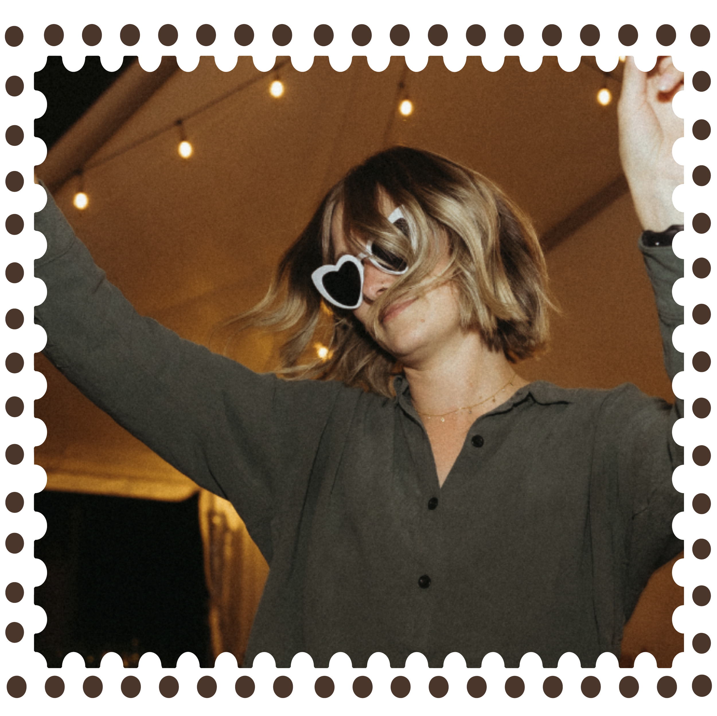 Person wearing heart-shaped sunglasses with blonde hair, smiling in front of a warm indoor background with string lights.