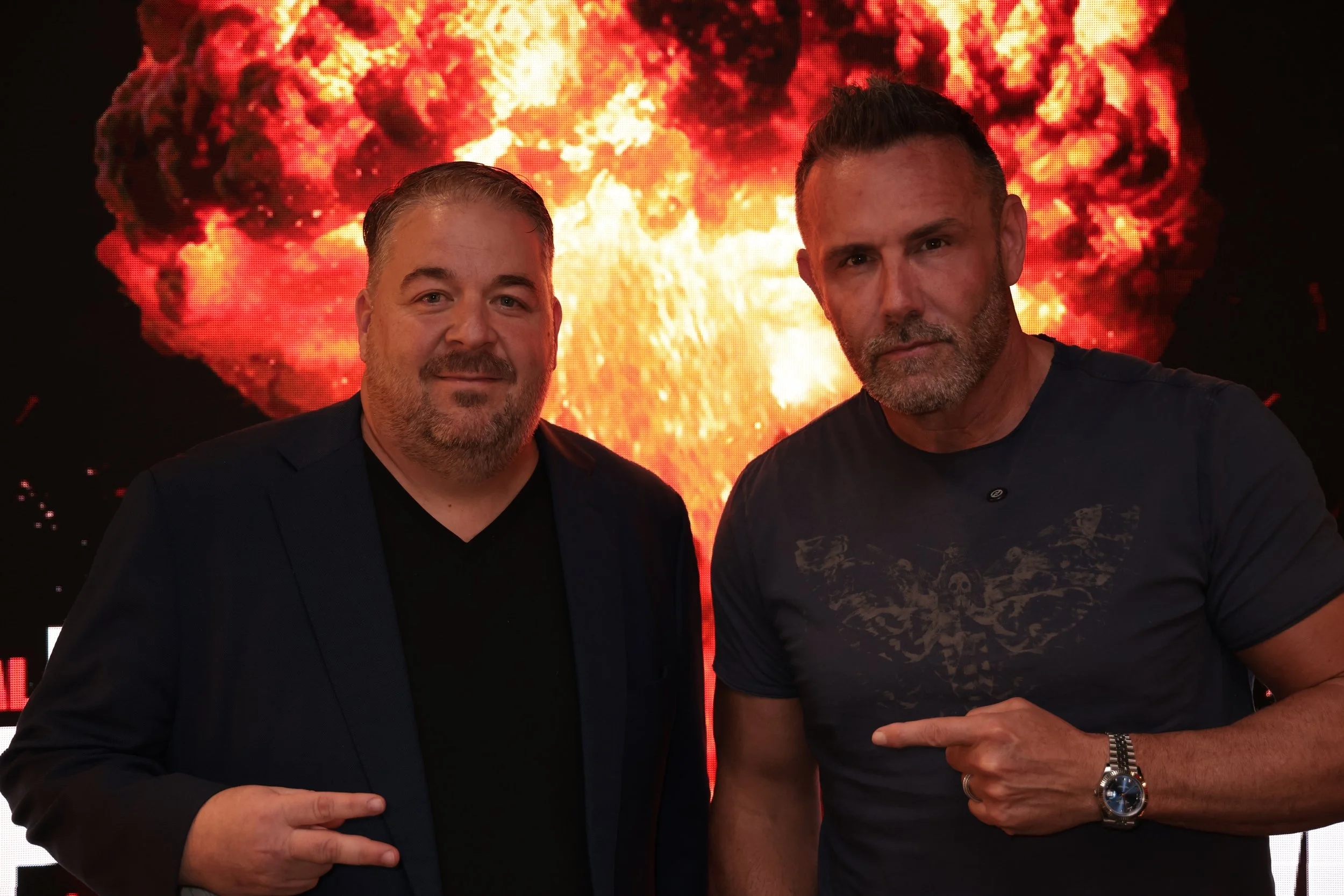 Two men stand in front of an explosive background. The man on the left has short, light-colored hair, a beard, and is wearing a dark blazer over a black shirt. The man on the right has short, gray hair and a beard, and is wearing a dark t-shirt with a skull and wings design, pointing to the man on the left with his right hand, wearing a watch and a bracelet.