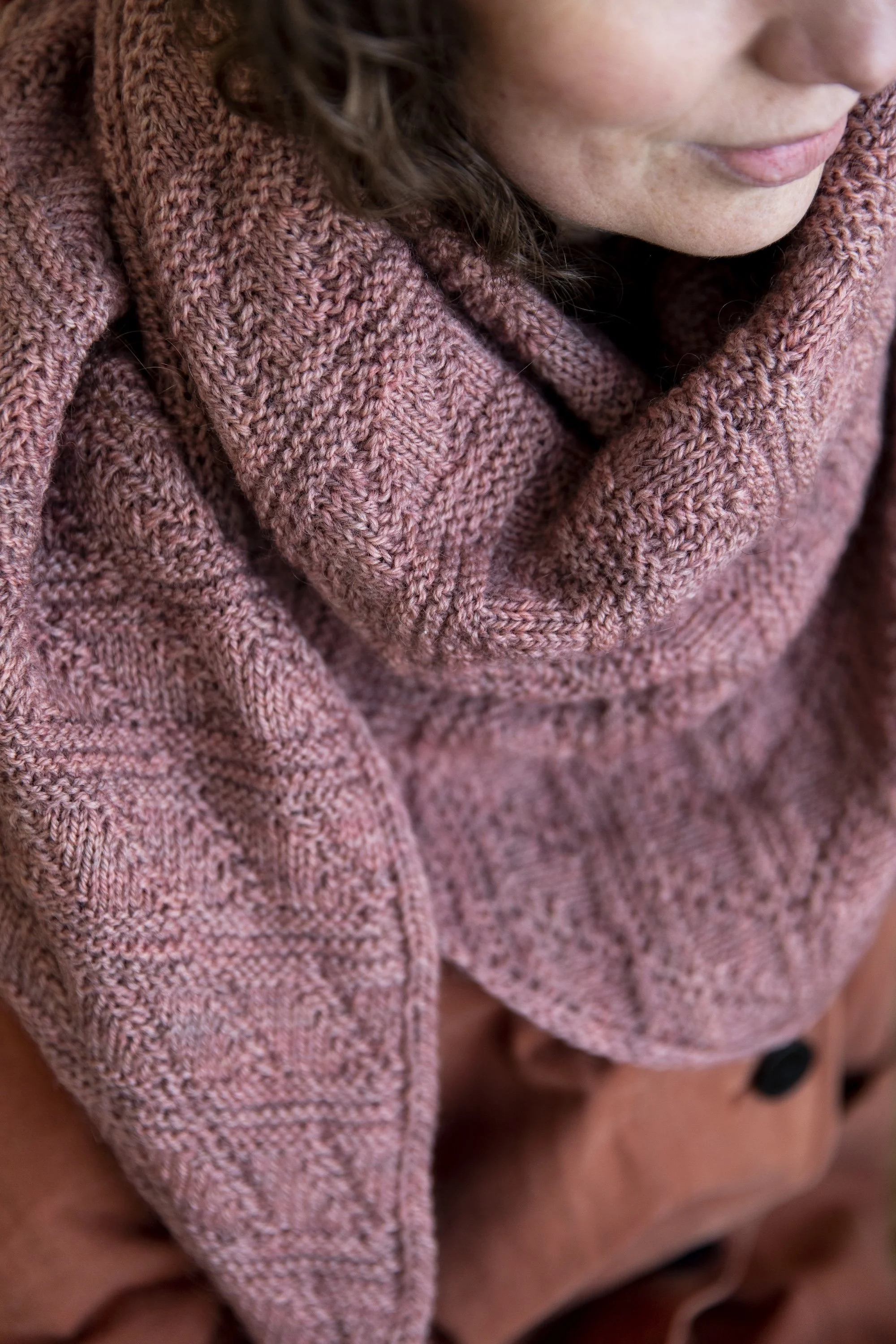 Close up of a hand-knitted shawl worn around the neck. The shawl is worked in dusty pink yarn, with a motif of diagonal lines crisscrossing vertical lines.