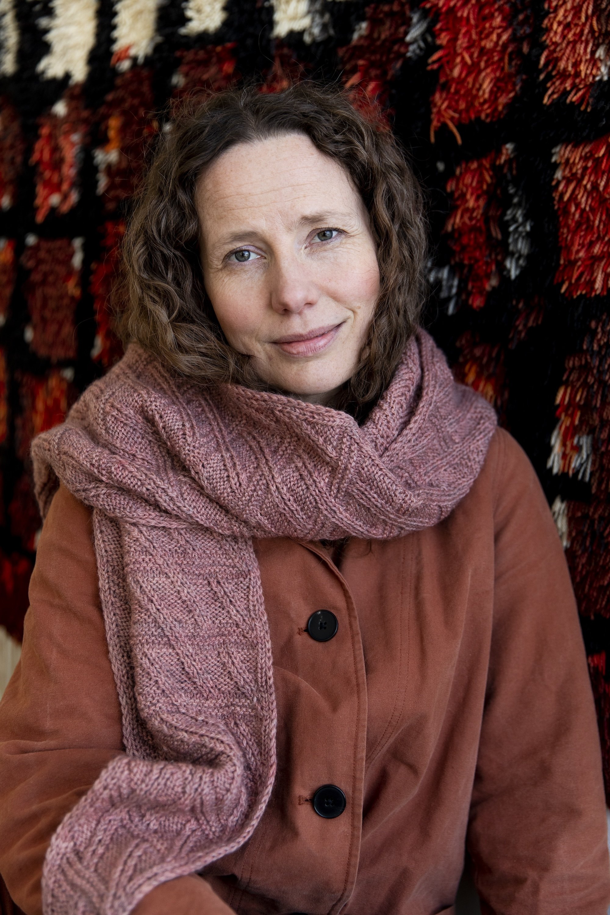 Model wearing a hand-knitted shawl around the neck over a buttoned up coat. The shawl is worked in dusty pink yarn, with a motif of diagonal lines crisscrossing vertical lines.