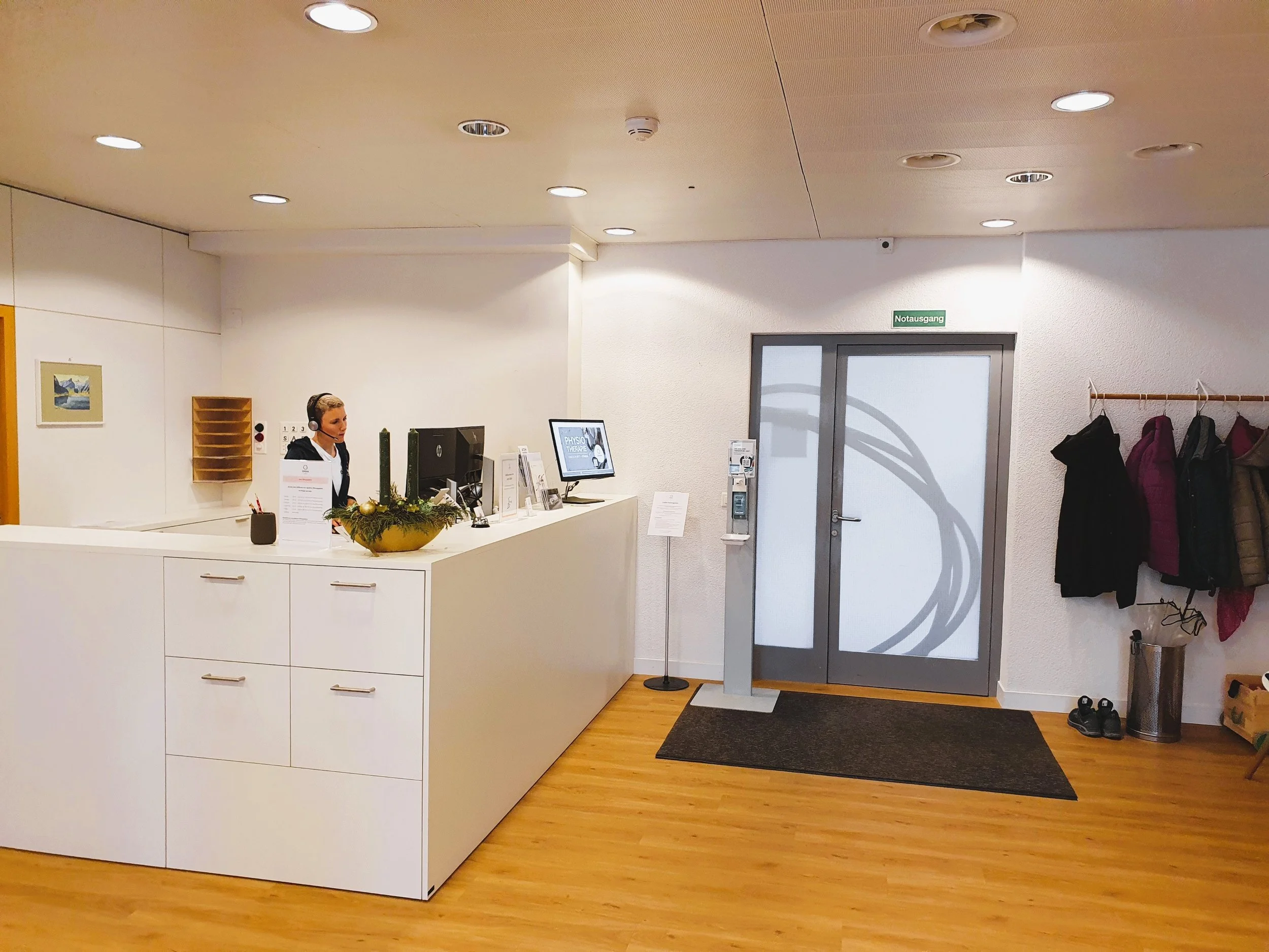 Rezeption mit einer weiblichen Angestellten mit Headset, gegenüber befindet sich ein Bildschirm, an der Wand rechts befindet sich eine Garderobe mit Mänteln, daneben ein Briefkasten, ein Handlauf und eine Tür mit einem Schild "Notausgang".