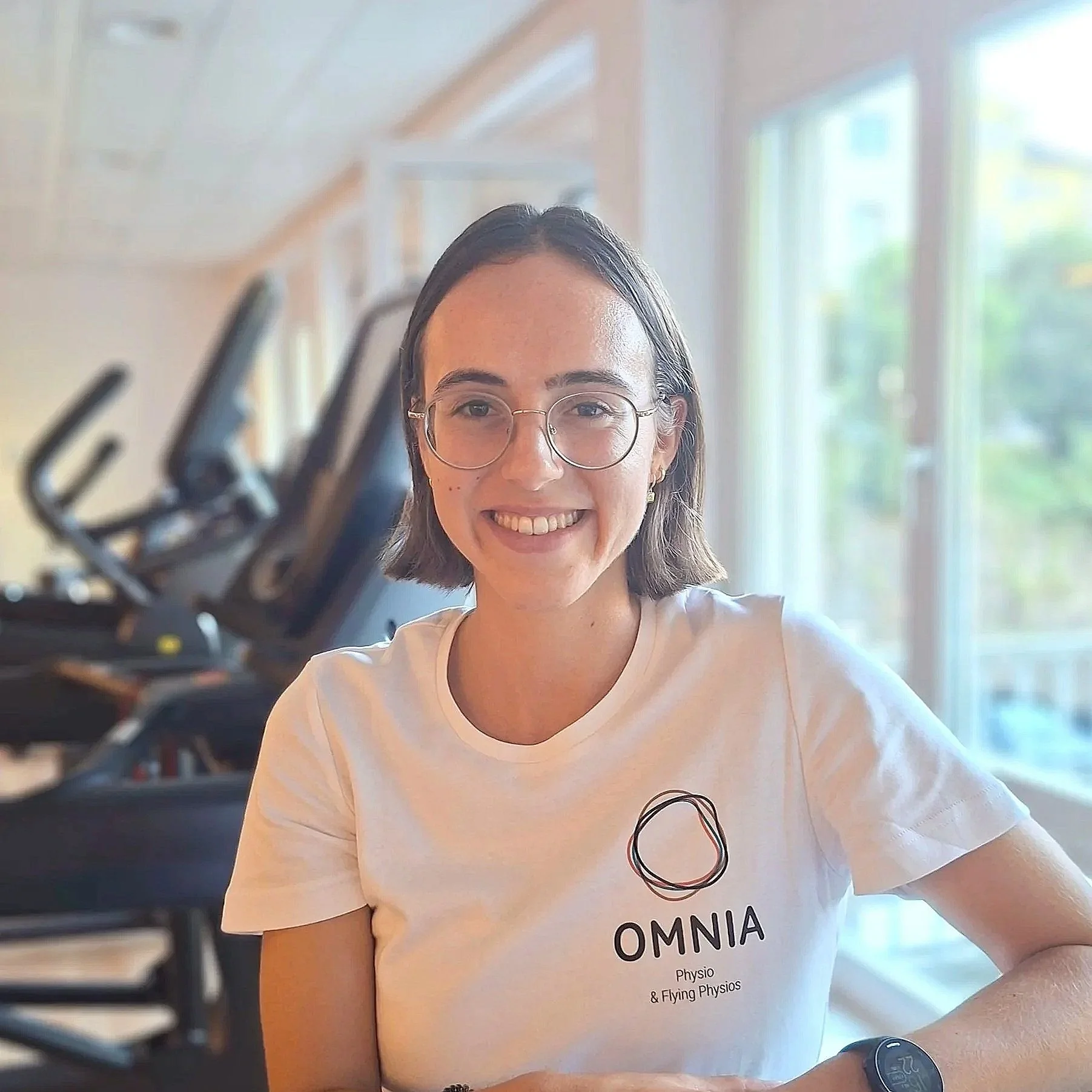 Junge Frau mit Brille und einem weißen T-Shirt, das das Wort 'OMNIA' und weitere Texte enthält, sitzt in einem Raum mit großen Fenstern, im Hintergrund ist ein Rack mit fünfen Radios. Sie lächelt und trägt einen schwarzen Fitness-Tracker an ihrem Handgelenk.