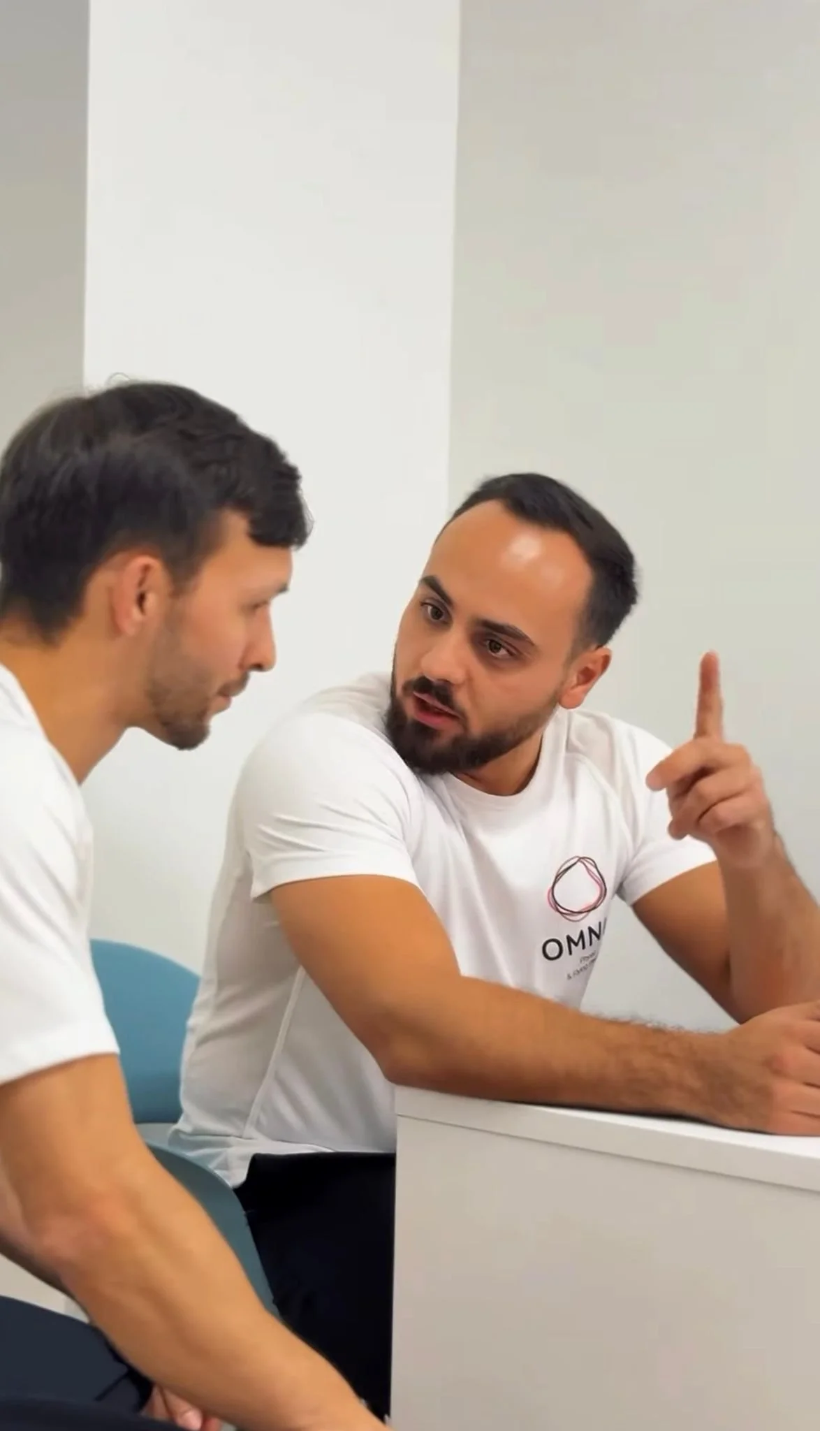 Zwei Männer im Gespräch, einer zeigt mit dem Finger nach oben, im Gespräch in einem Raum mit weißer Wand, sie tragen weiße T-Shirts.