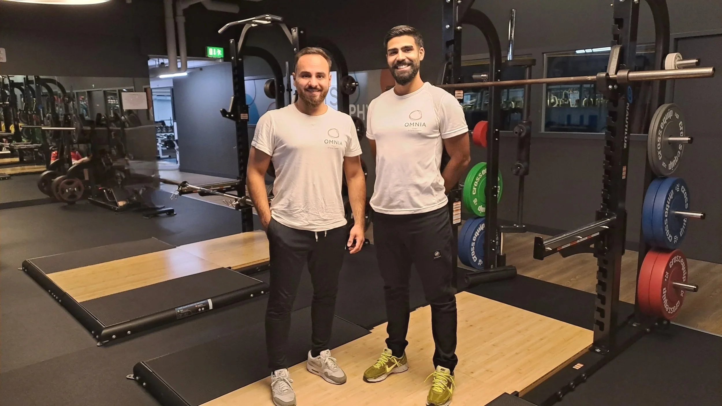 Zwei Männer stehen in einem Fitnessstudio vor einer Kraftstation, beide tragen weiße T-Shirts mit dem Logo 'OMNIA', lächeln und schauen in die Kamera.