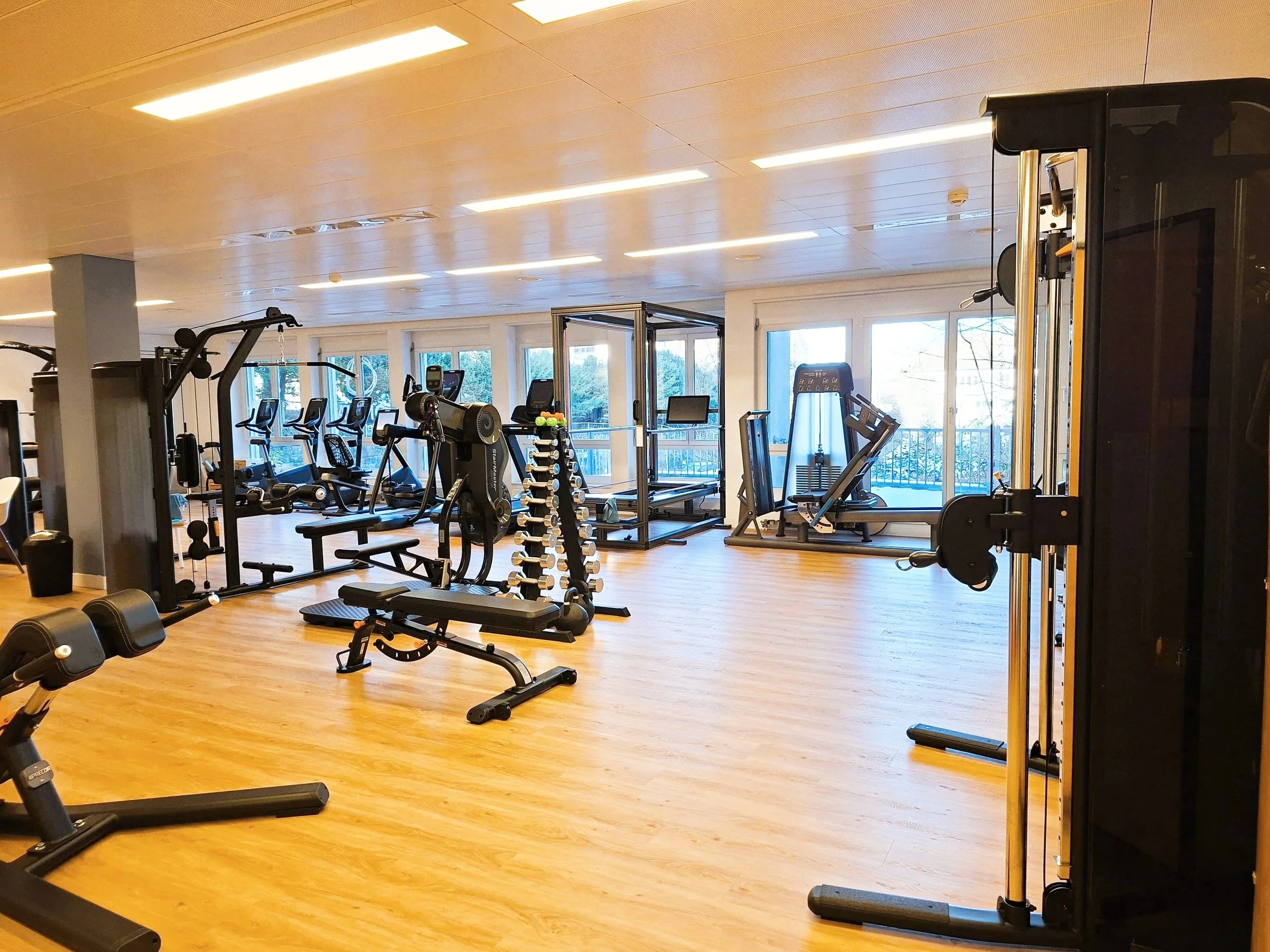 Leerer Fitnessraum mit verschiedenen Trainingsgeräten wie Hanteln, Laufbändern und Kraftmaschinen, große Fenster mit Aussicht auf die Natur.