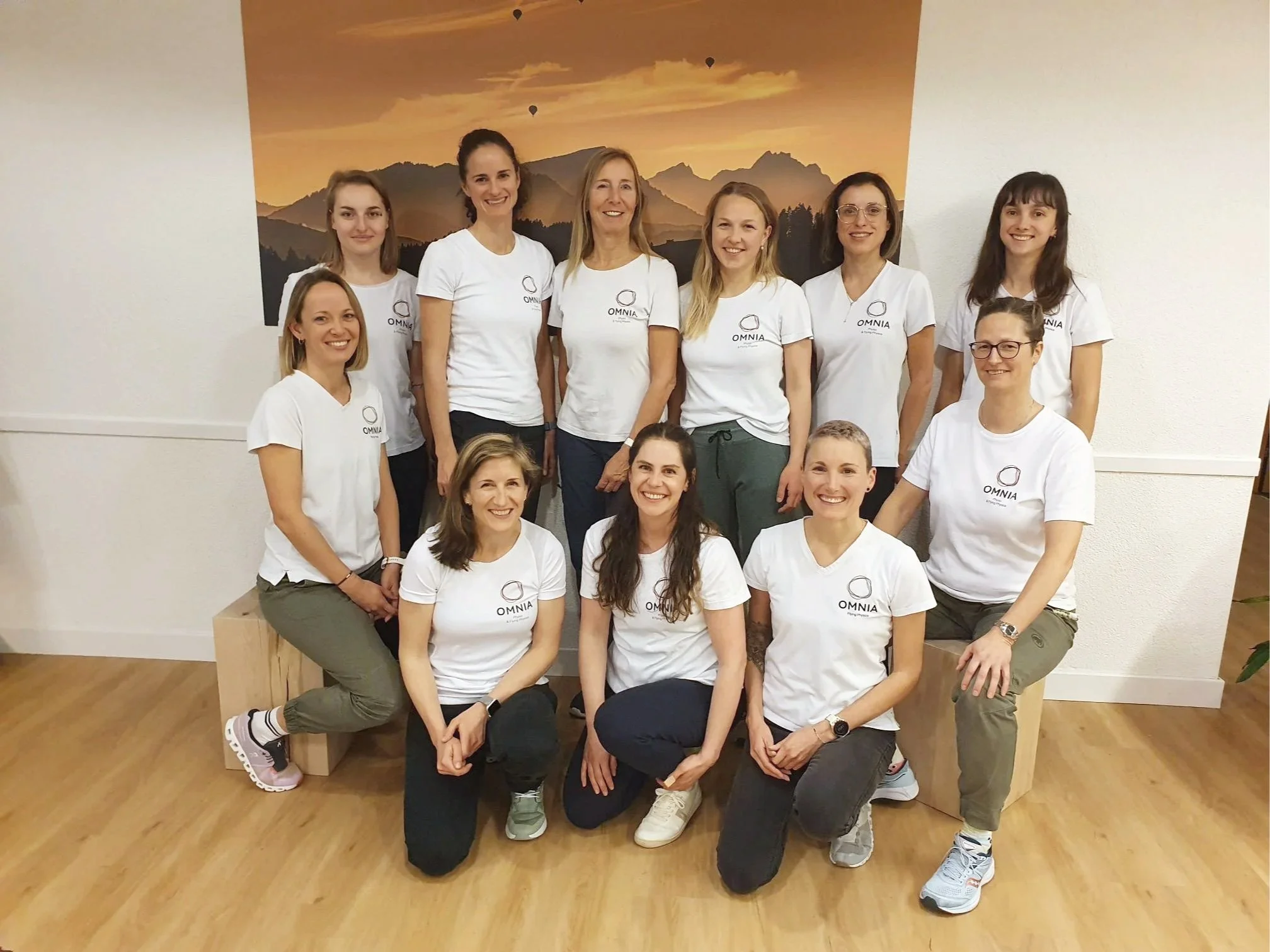 Gruppe von 12 Frauen, die alle weiße T-Shirts mit dem Logo 'OMNIA' tragen, vor einer Wand mit Berglandschaft und Heißluftballons. Sie posieren für ein Gruppenbild in einem Raum mit Holzboden.