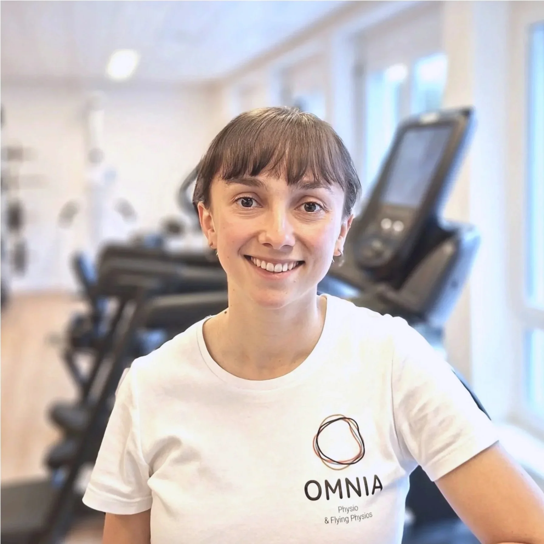 Junge Frau mit Kurzhaar und weißen T-Shirt mit OMNIA-Logo im Fitnessstudio, unscharfe Trainingsgeräte im Hintergrund.