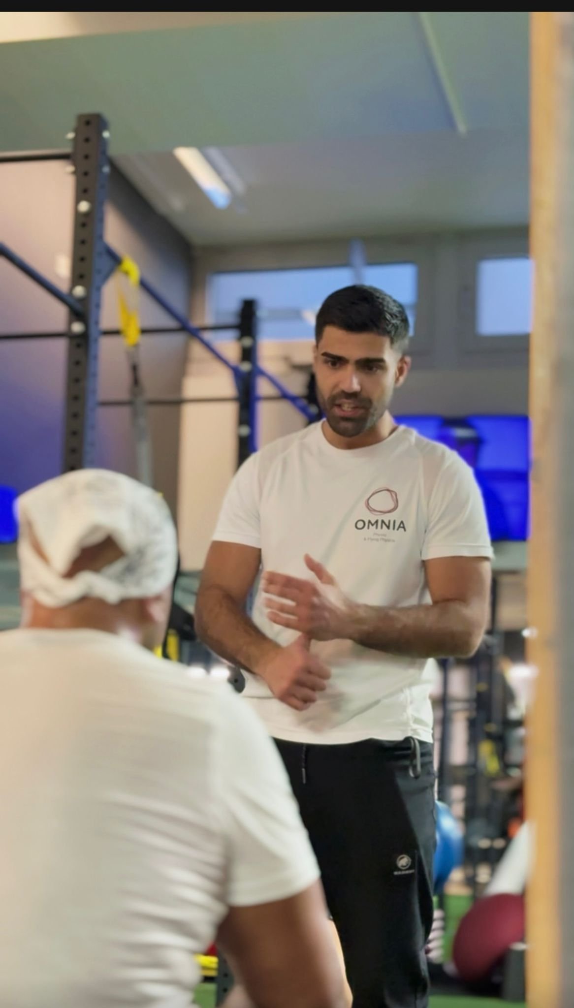 Ein Mann erklärt einer Person im Fitnessstudio. Im Hintergrund Trainingsgeräte und Fitnessequipment.