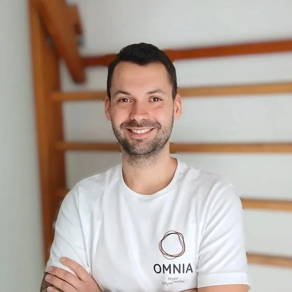 Ein lachender Mann trägt ein weißes T-Shirt mit dem Logo und dem Text 'OMNIA Physio & Flying Physiotherapy'. Er steht vor einer Holzwand.