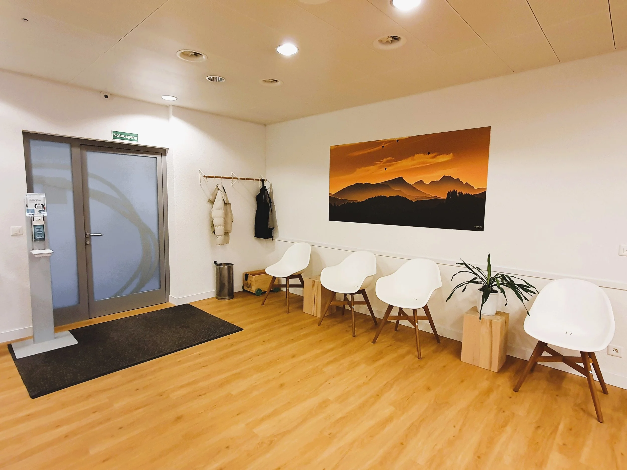 Wartezimmer mit weißen Stühlen, einem Bild von Bergen bei Sonnenuntergang, mehreren Jacken an einer Garderobe, einem Pflanzenkübel und einem Eingang mit Notausgangsschilder.