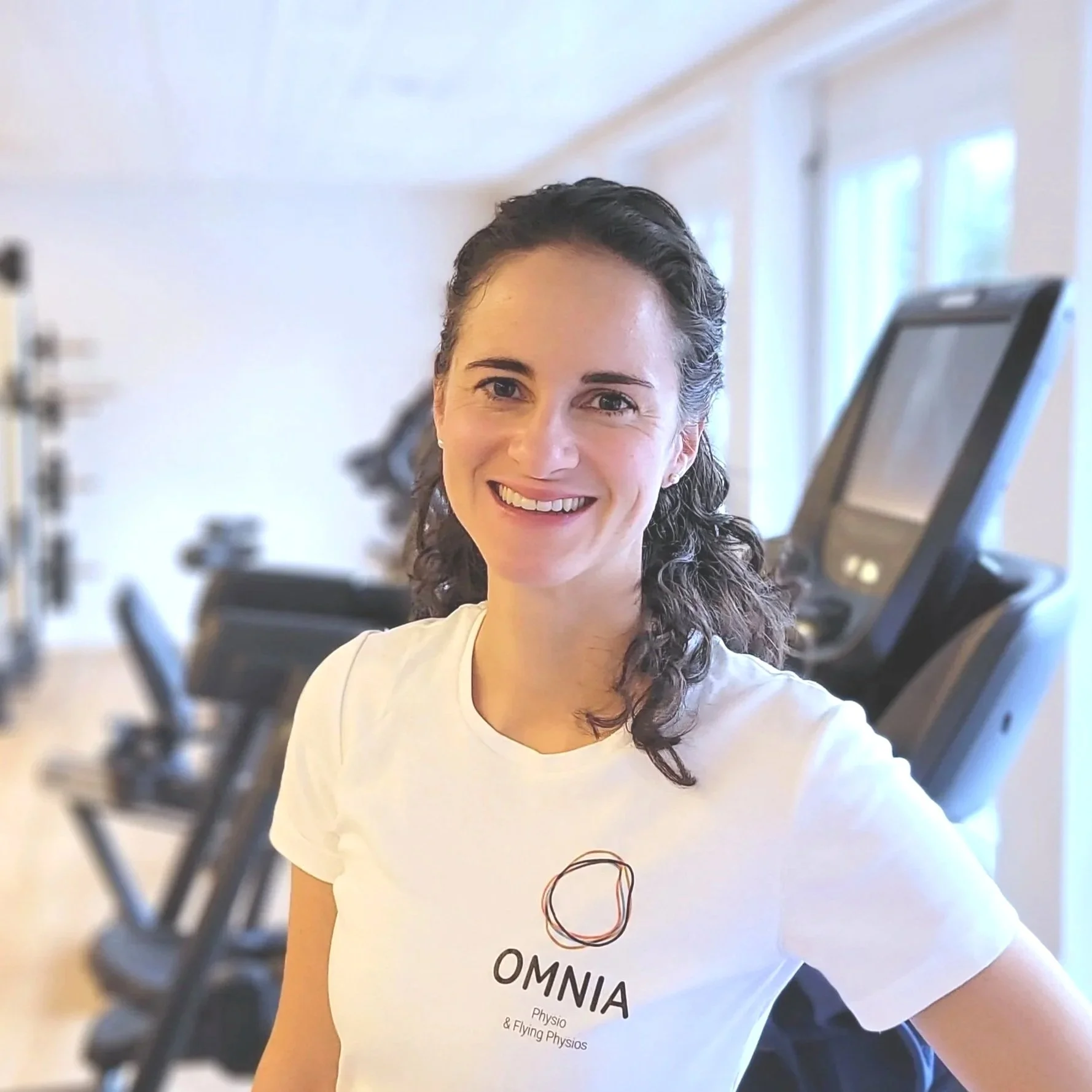 Junge Frau in Fitnessstudio, lacht, trägt ein weißes T-Shirt mit OMNIA-Logo, im Hintergrund Laufbänder.