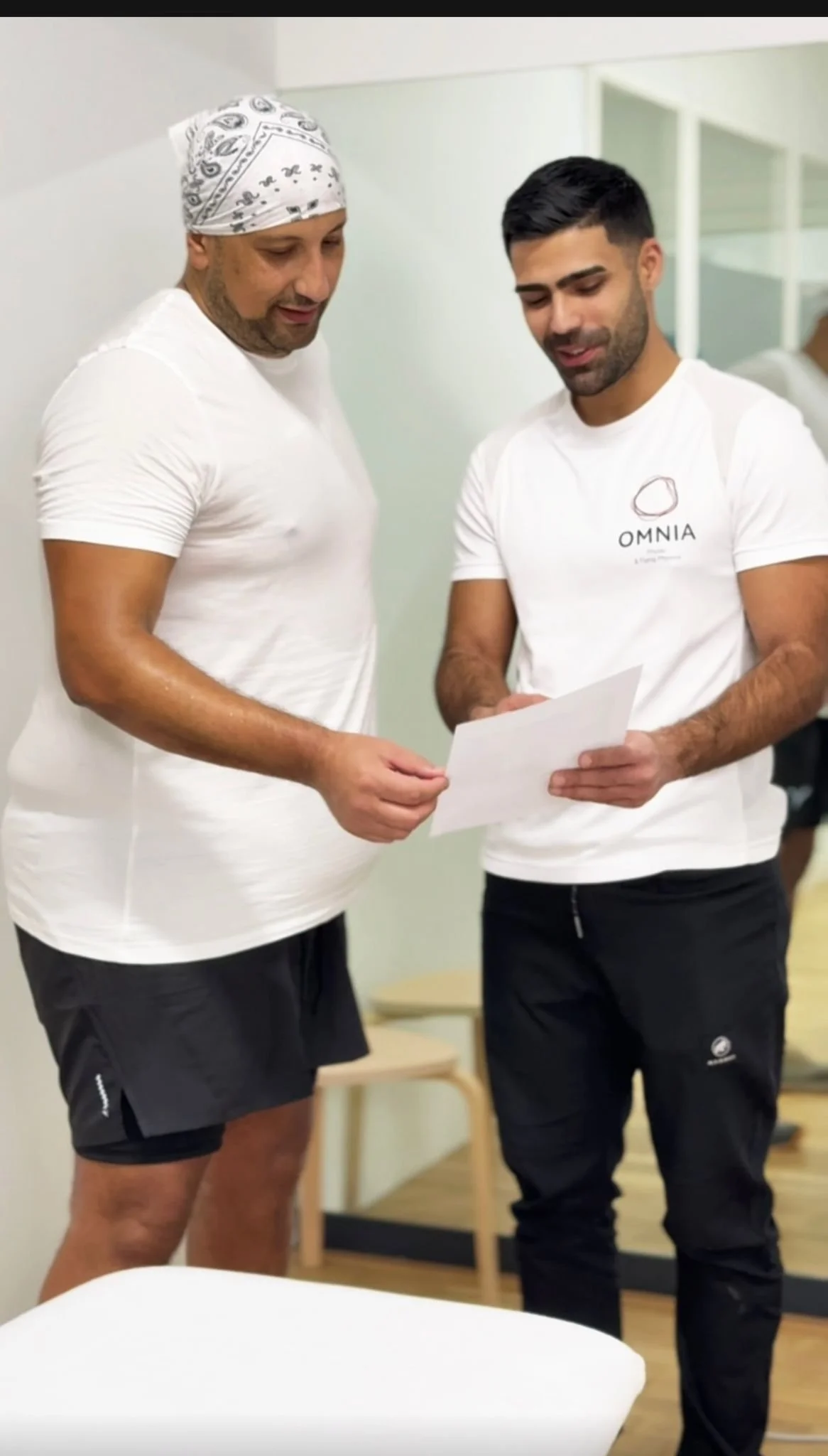 Zwei Männer schauen gemeinsam auf ein Dokument, einer trägt ein weißes T-Shirt mit der Aufschrift 'OMNIA' und der andere trägt ein Bandana. Sie stehen in einem Raum mit Spiegeln.